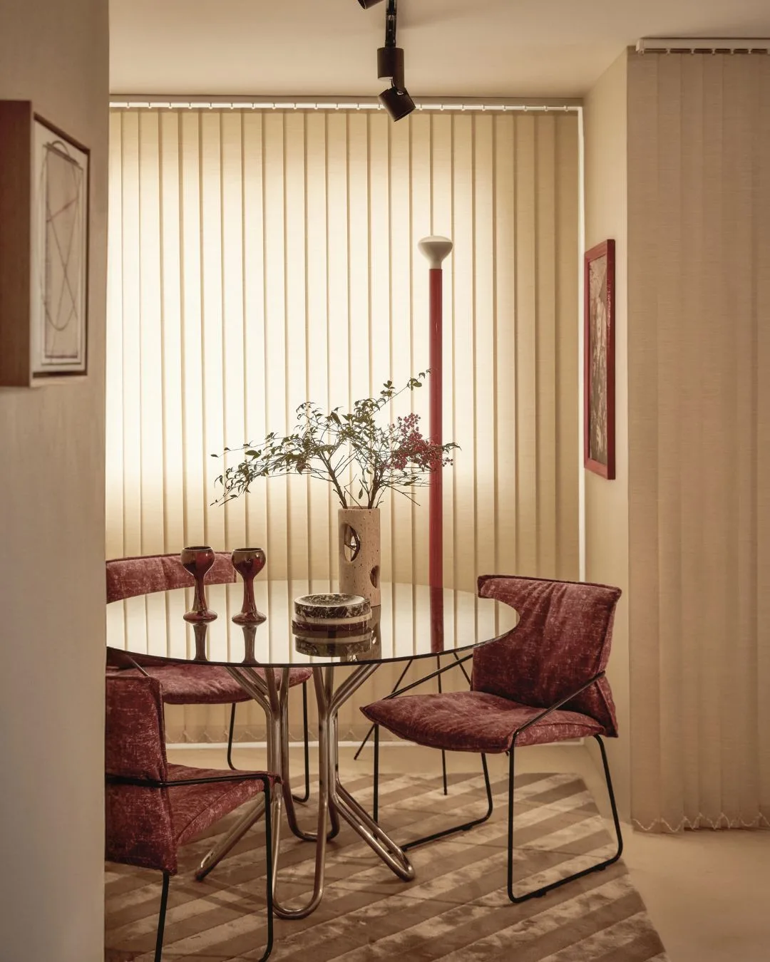 A dining area with a glass table, red-pink chairs, ambient lighting, vertical blinds, and decorative vessels.