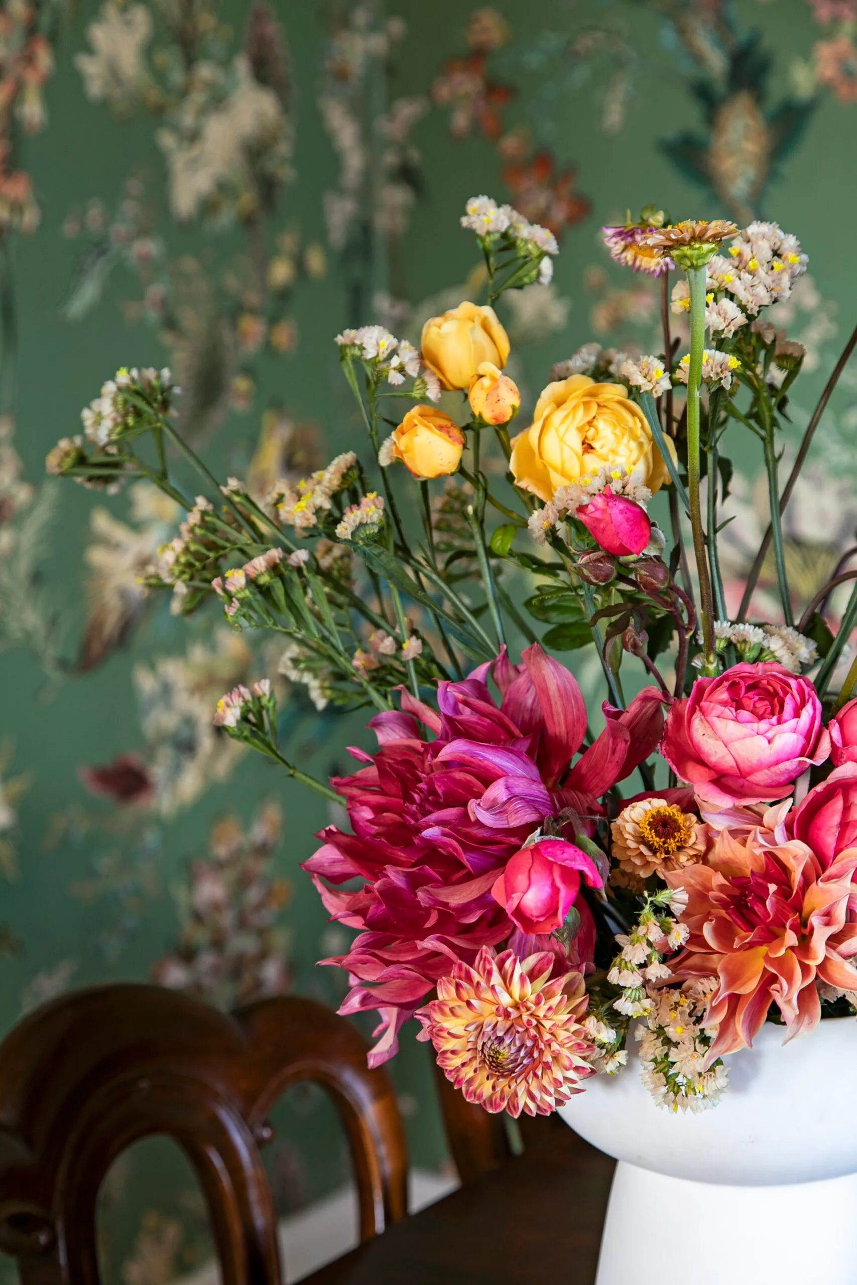 A vibrant bouquet in a white vase in front of green floral wallpaper.