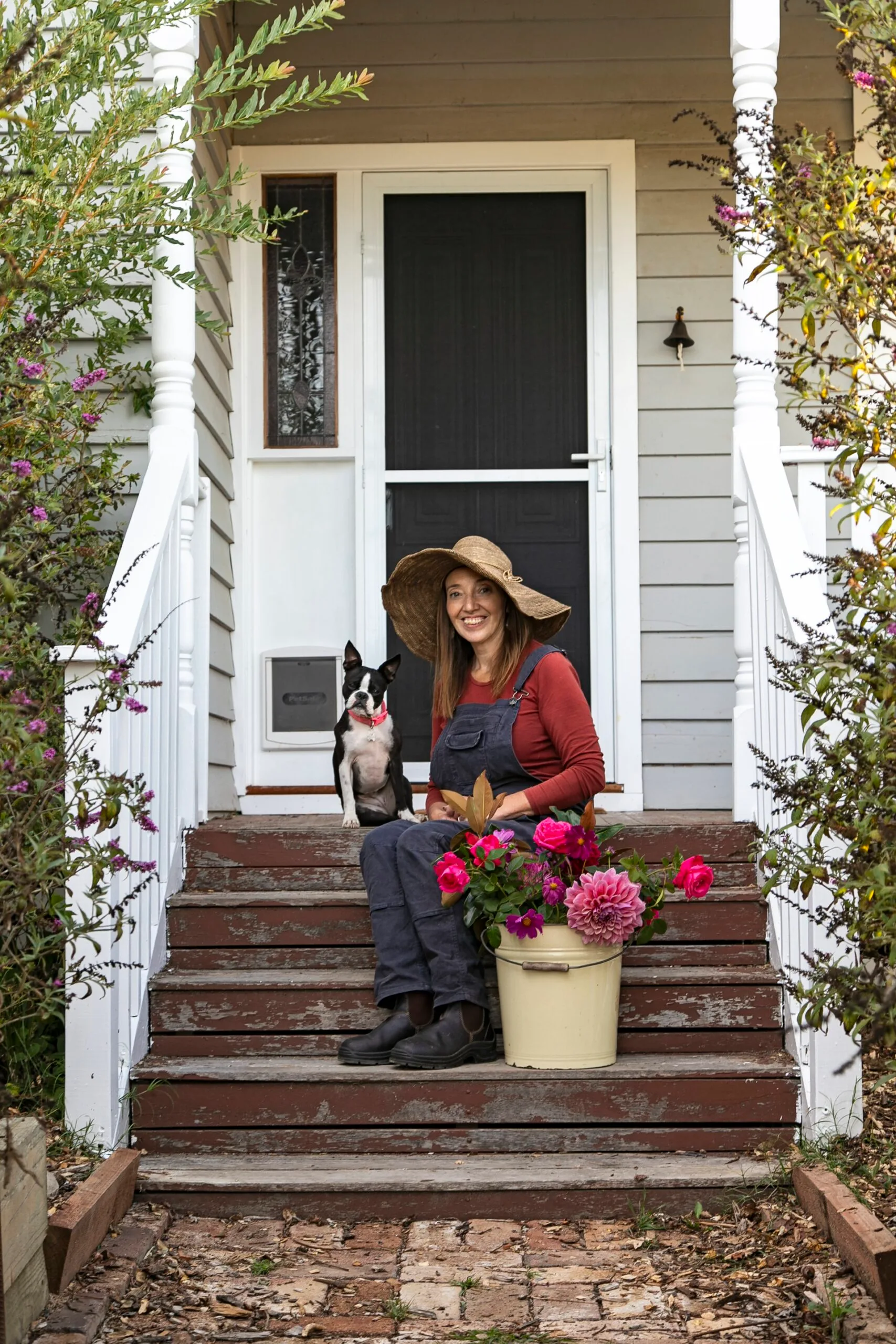 A woman on the front steps of a country cottage with her Boston Terrier dog.