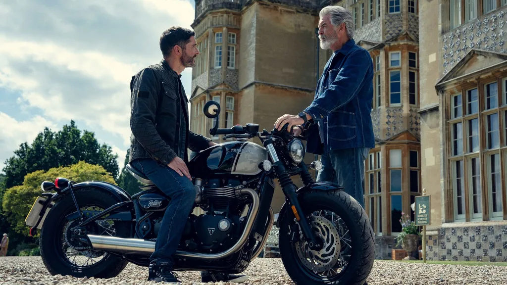 An older man wearing a denim outfit speaks to a younger man sitting on a motorbike outside a stately home in The Thursday Murder Club Netflix film.