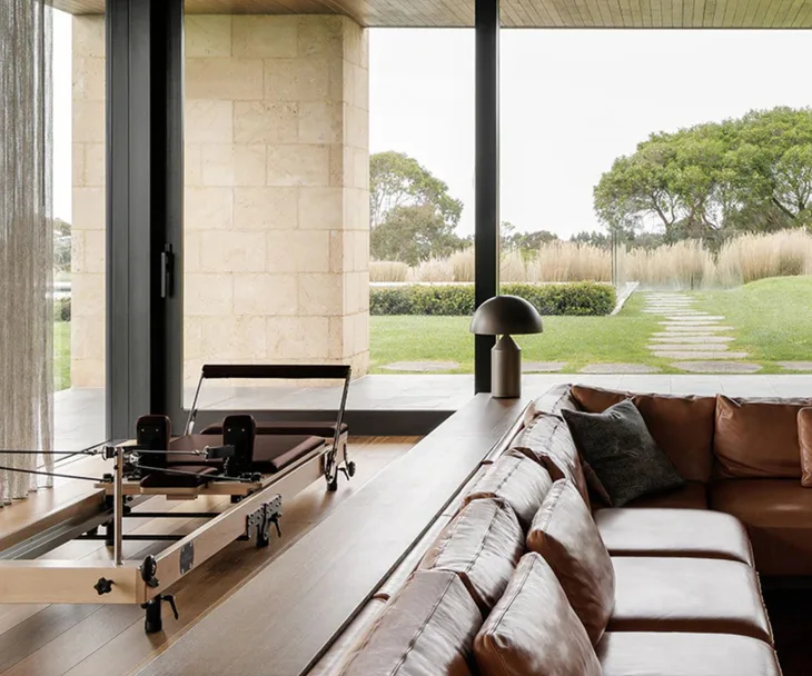 A brown coloured pilates reformer machine in an architectural living room surrounded by large windows and a sunken brown leather lounge area.