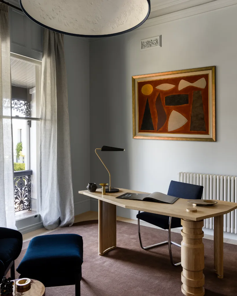 Modern office interior with a wooden desk, abstract painting, and a large window with sheer curtains. Mim Design verdant residence