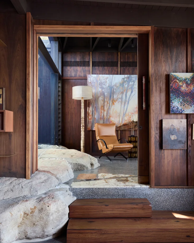 Cozy wooden interior with a tan leather chair, abstract wall art, and a tall lamp, featuring natural stone elements.