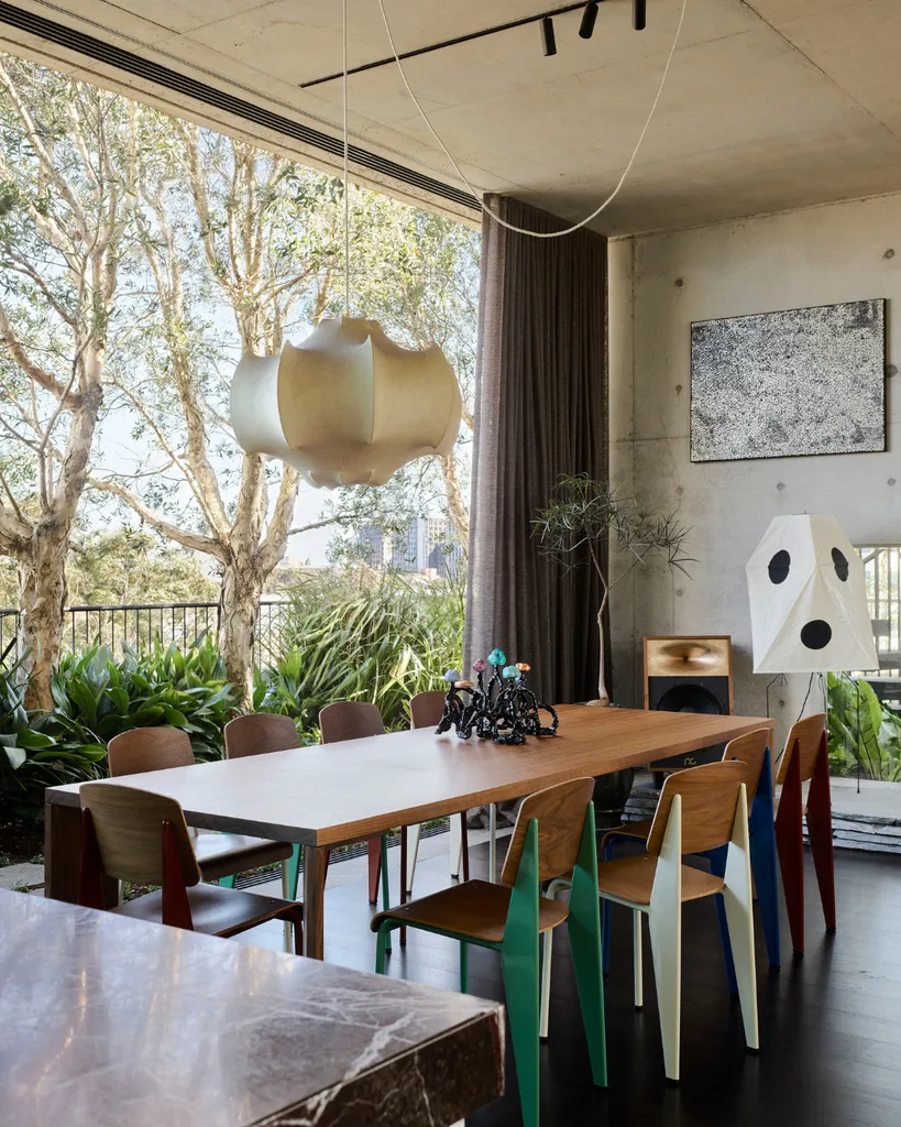 Dining room with a wooden table, colorful chairs, sculptural pendant light, and large window overlooking greenery.