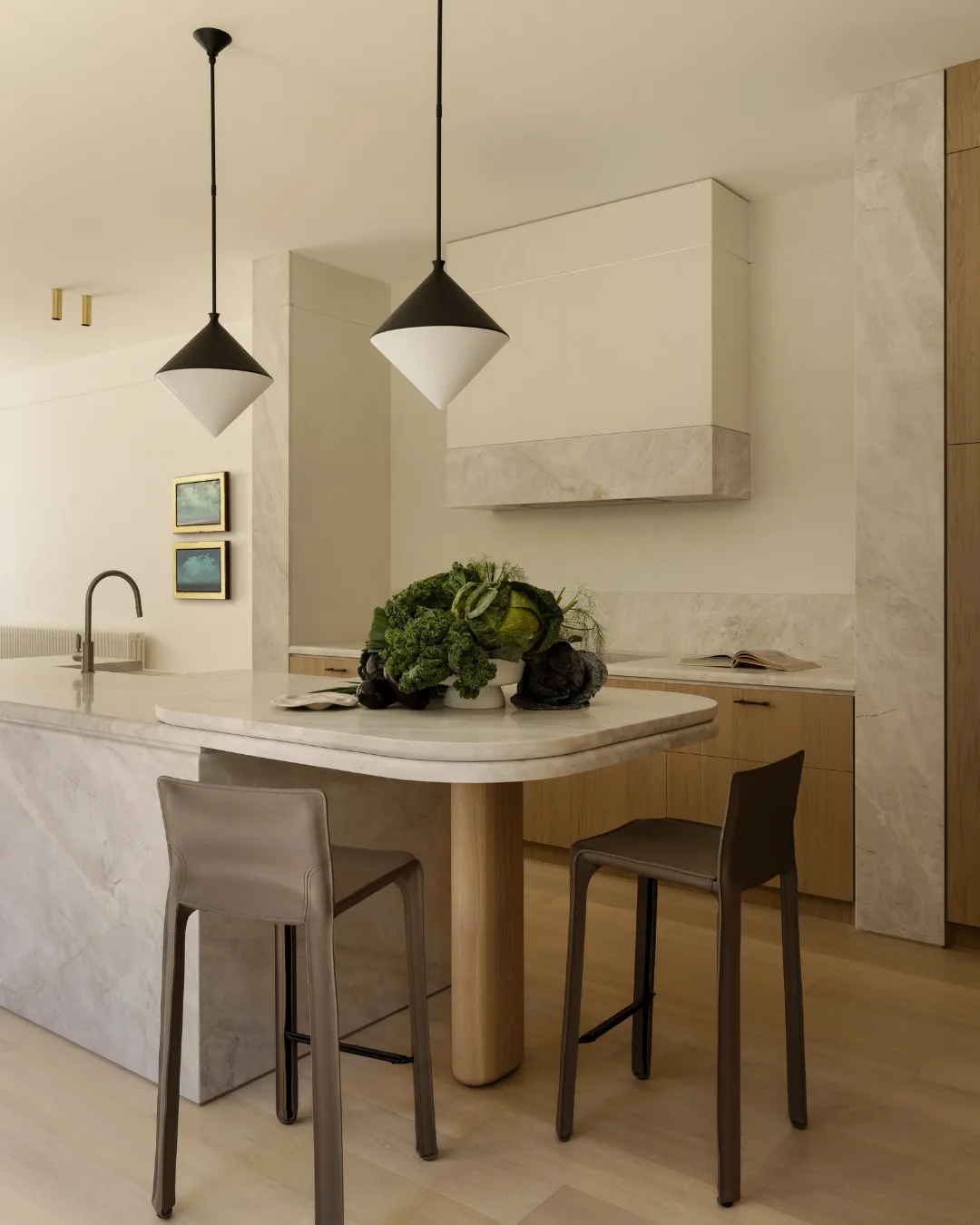 Kitchen with marble island, two pendant lights, two brown stools, and a bowl of vegetables. Minimalist decor. Mim Design verdant residence.
