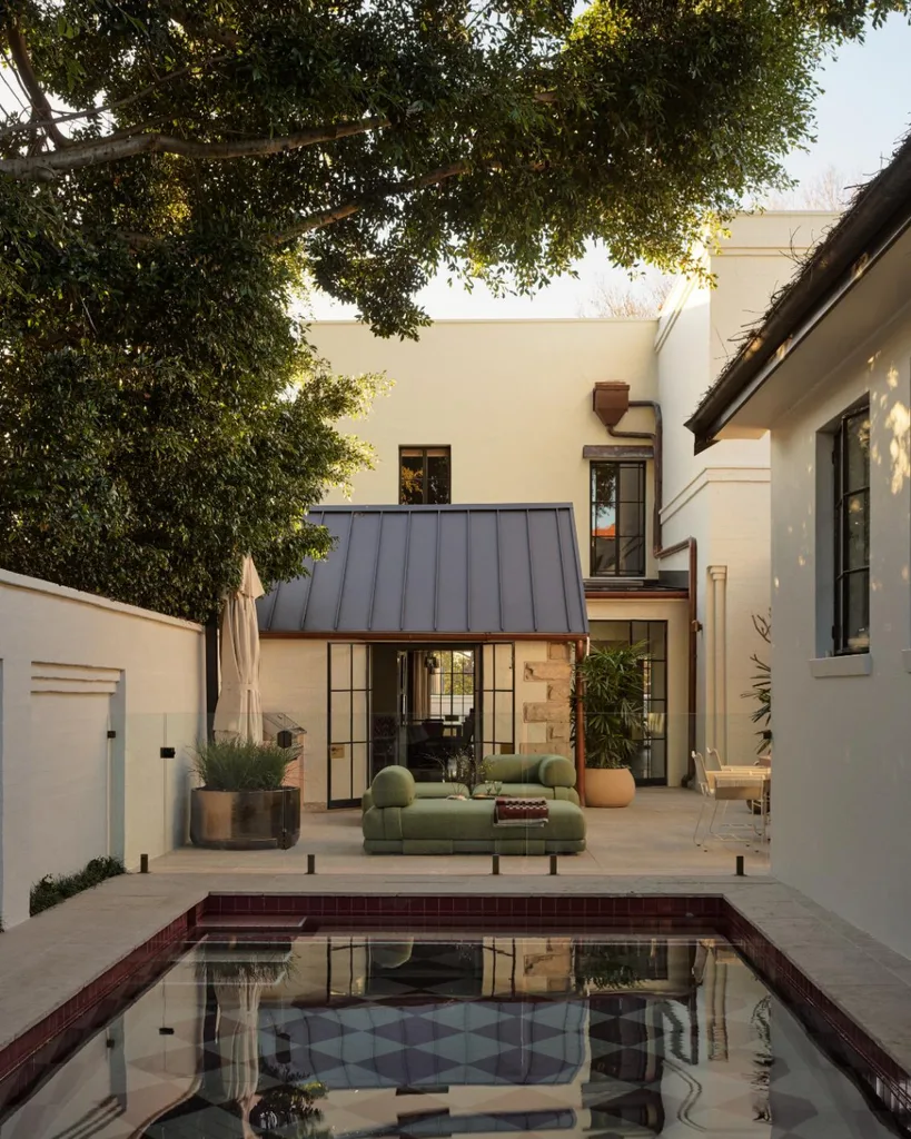 Contemporary patio with green sofa, pool, and shaded by trees, featuring a modern building with large windows in the background.