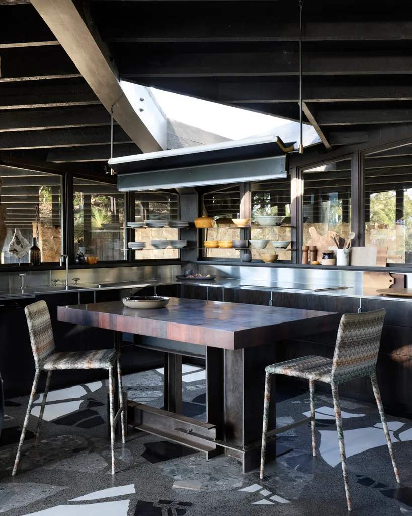 A modern kitchen with a large wooden table, two patterned chairs, and glass shelves with bowls.