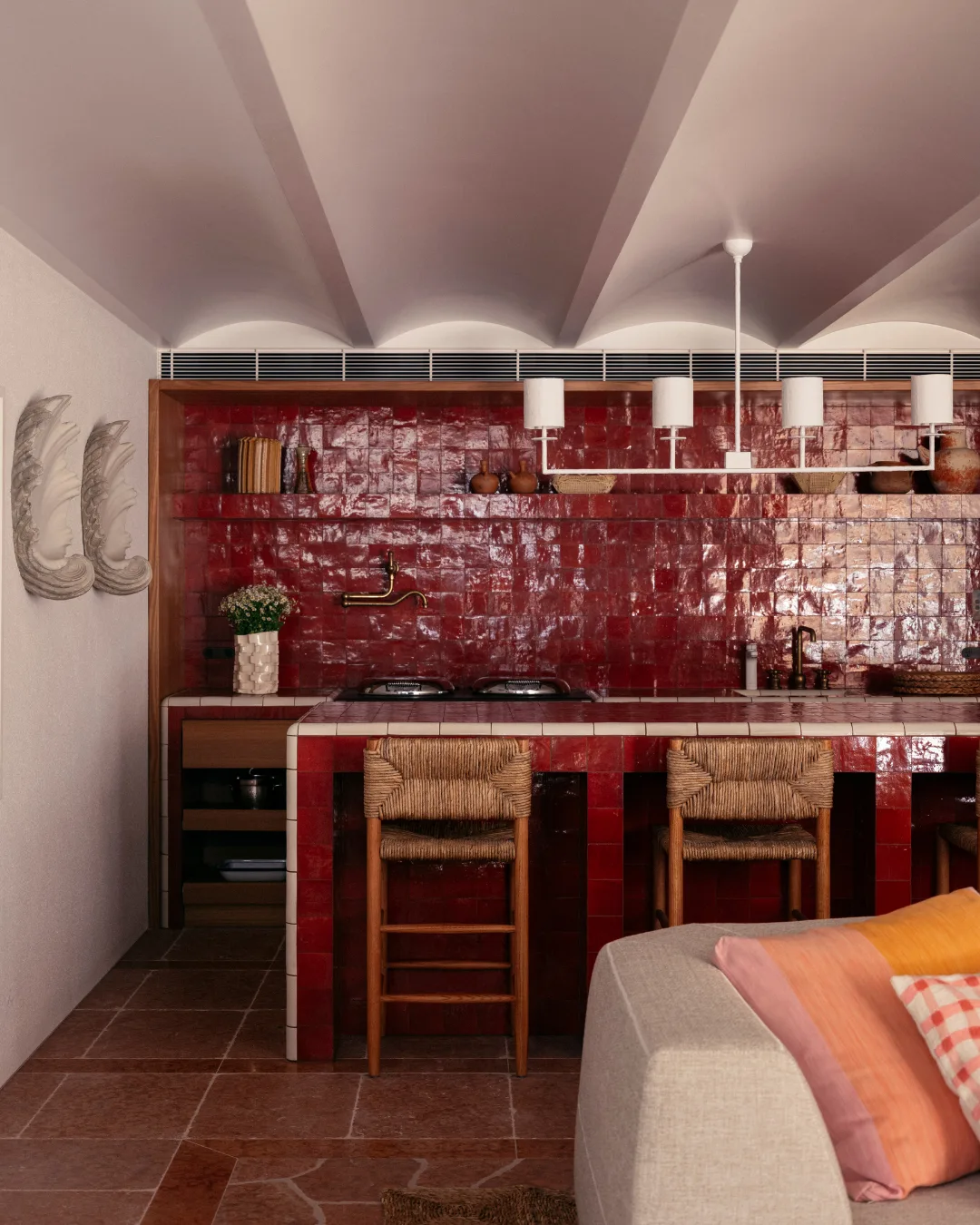 Red-tiled kitchen with wicker chairs, modern chandelier, decorations, and a beige sofa with colorful pillows.