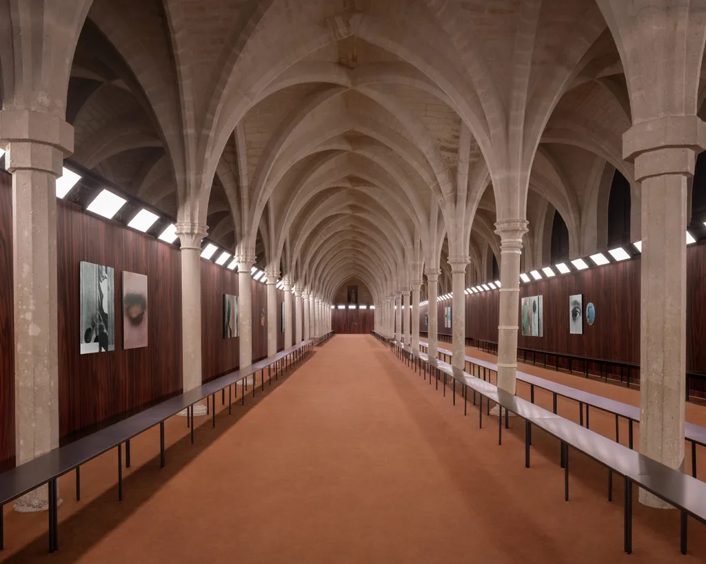 Elegant hall with stone arches, lined with artwork on wooden walls, and illuminated by bright, evenly spaced lights. |