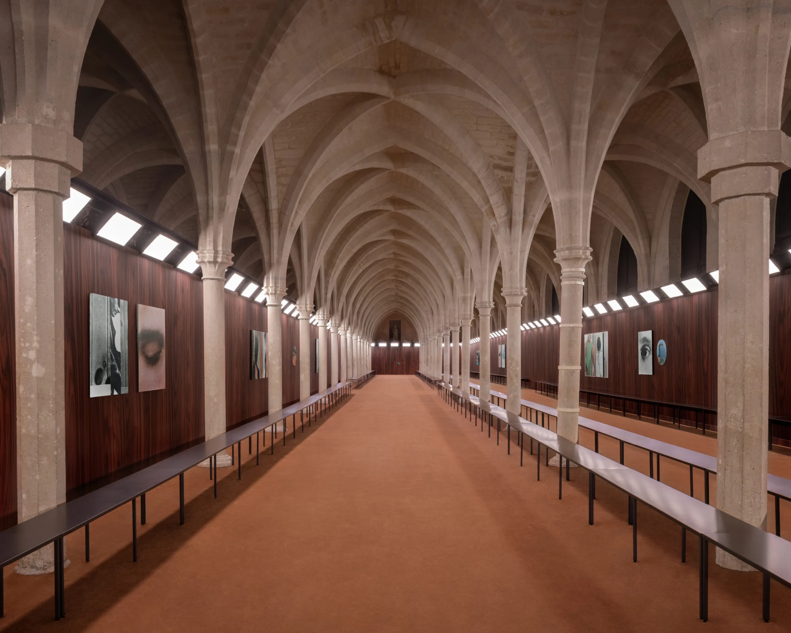 Elegant hall with stone arches, lined with artwork on wooden walls, and illuminated by bright, evenly spaced lights. |