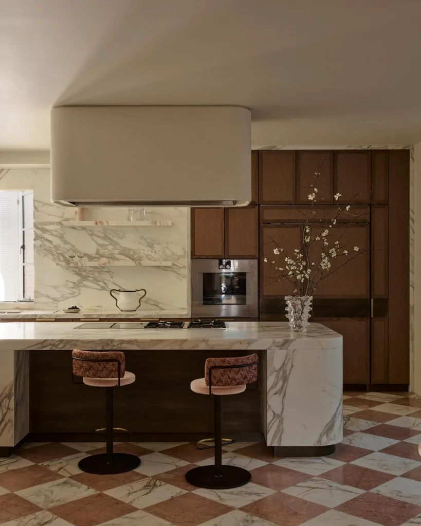 Luxurious kitchen with marble countertops, two pink bar stools, wood cabinets, and a vase with branches on the island.