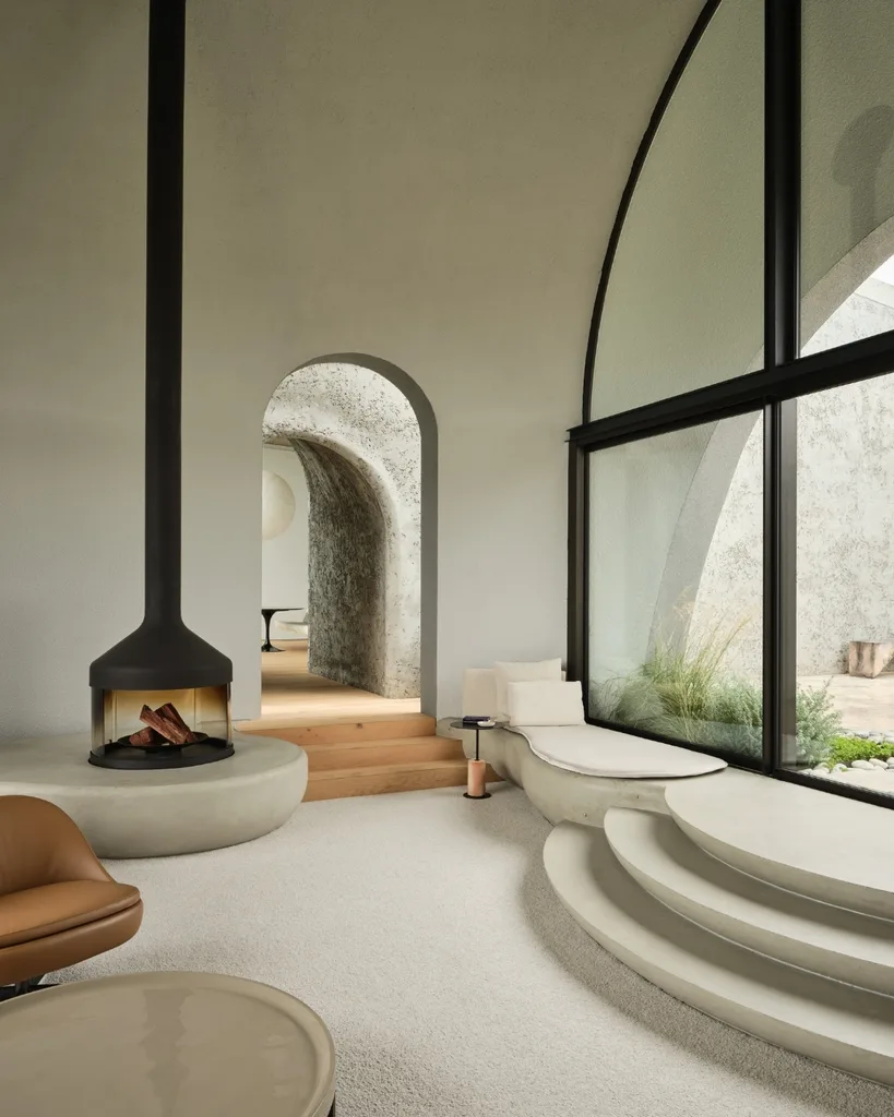 Modern living area with arched window, curvy built-in seating, central fireplace, and wooden steps leading to another room.
