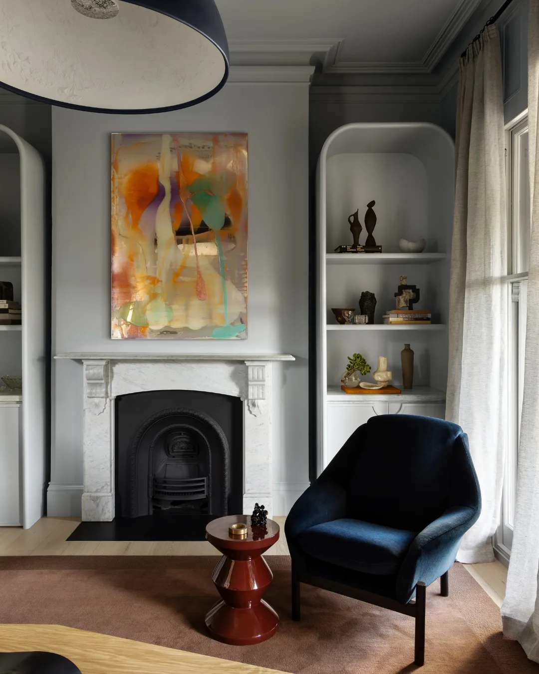 Stylish living room with abstract art, a marble fireplace, blue armchair, and curved shelves displaying decor items. Mim Design verdant residence.