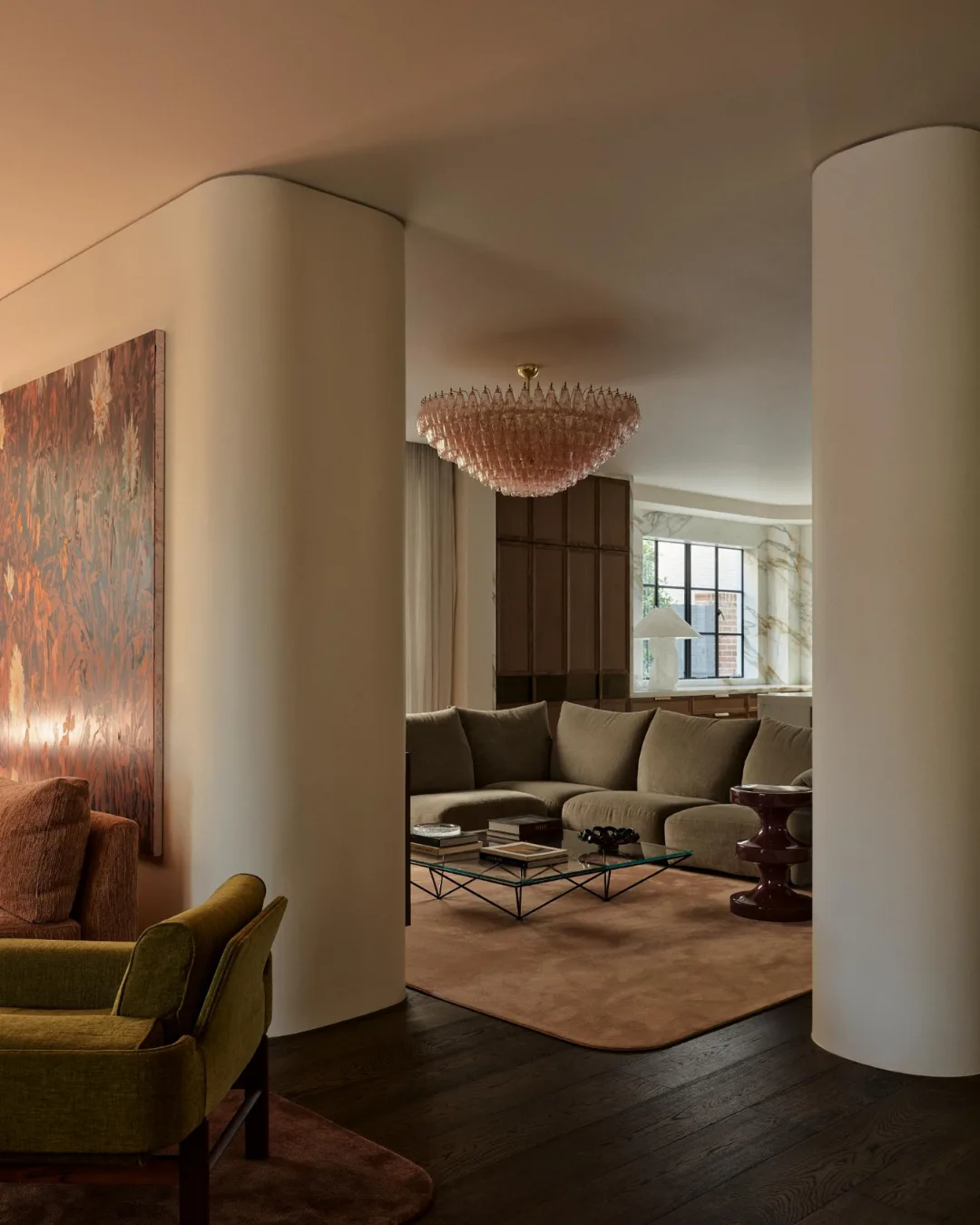 Luxurious living room with a beige sectional sofa, glass coffee table, artwork, and a pink chandelier.