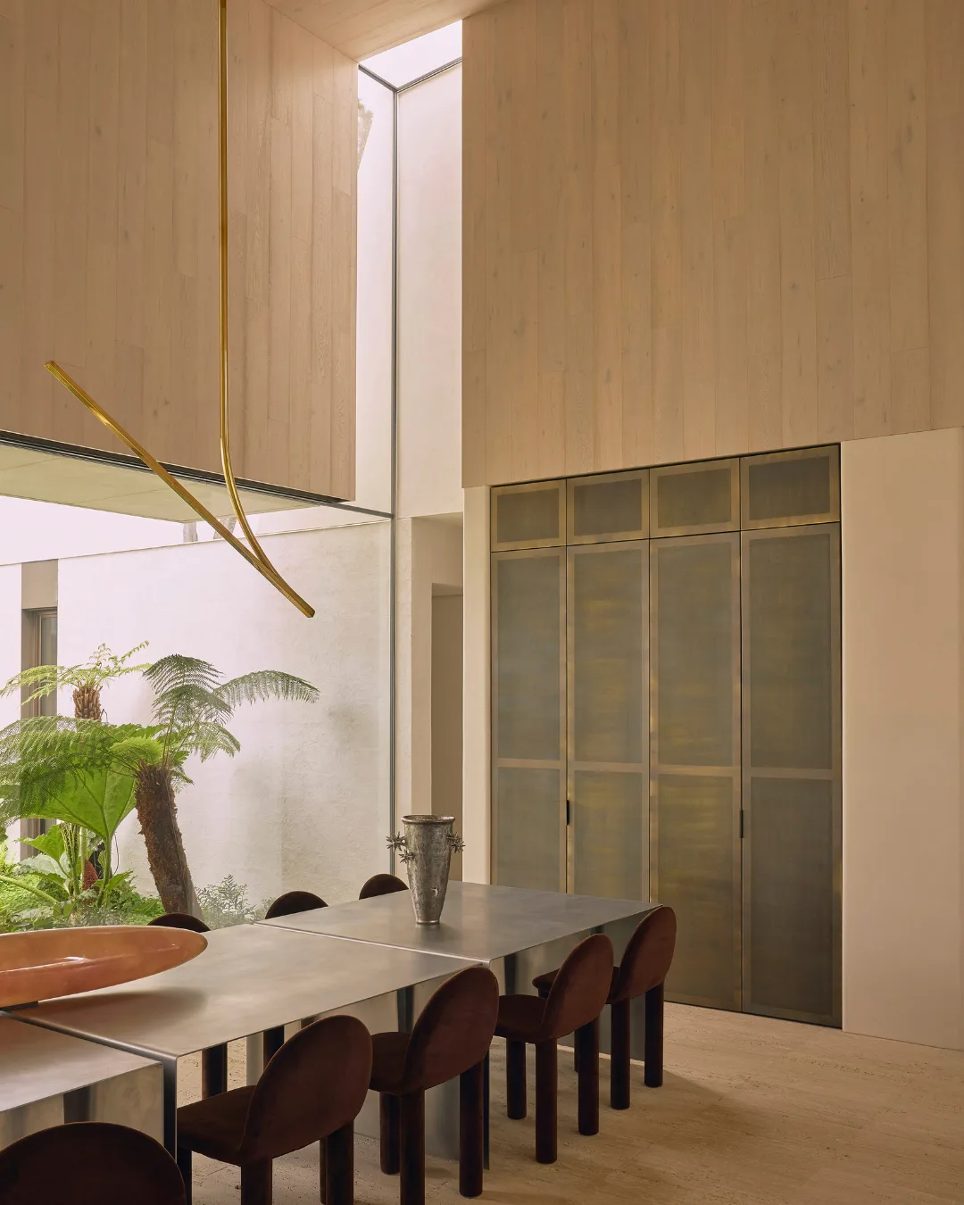 Modern dining room with a long table, dark chairs, high ceiling, large windows, and a view of greenery outside.