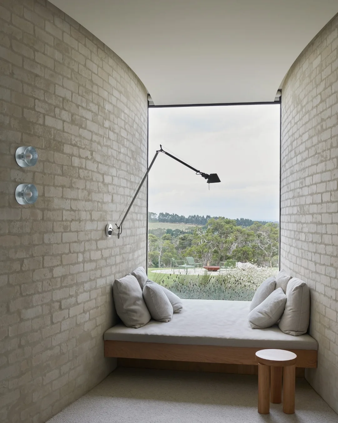 Cozy nook with beige cushions, large window view of garden, and modern adjustable wall lamp. 