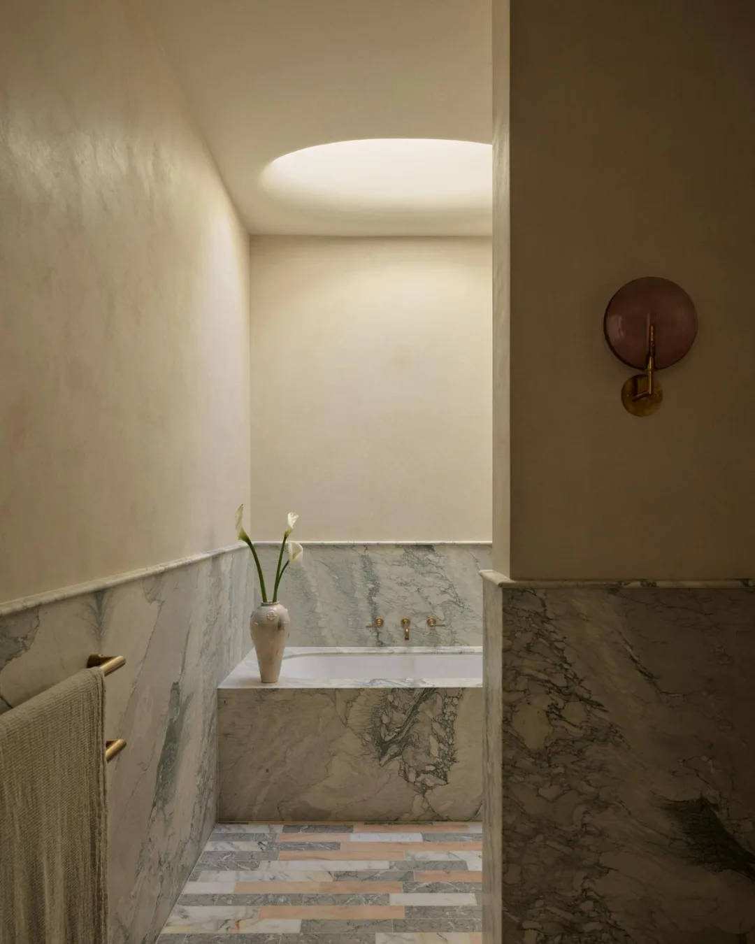 Luxurious bathroom with marble tub, vase with flowers, and soft lighting from a circular skylight.