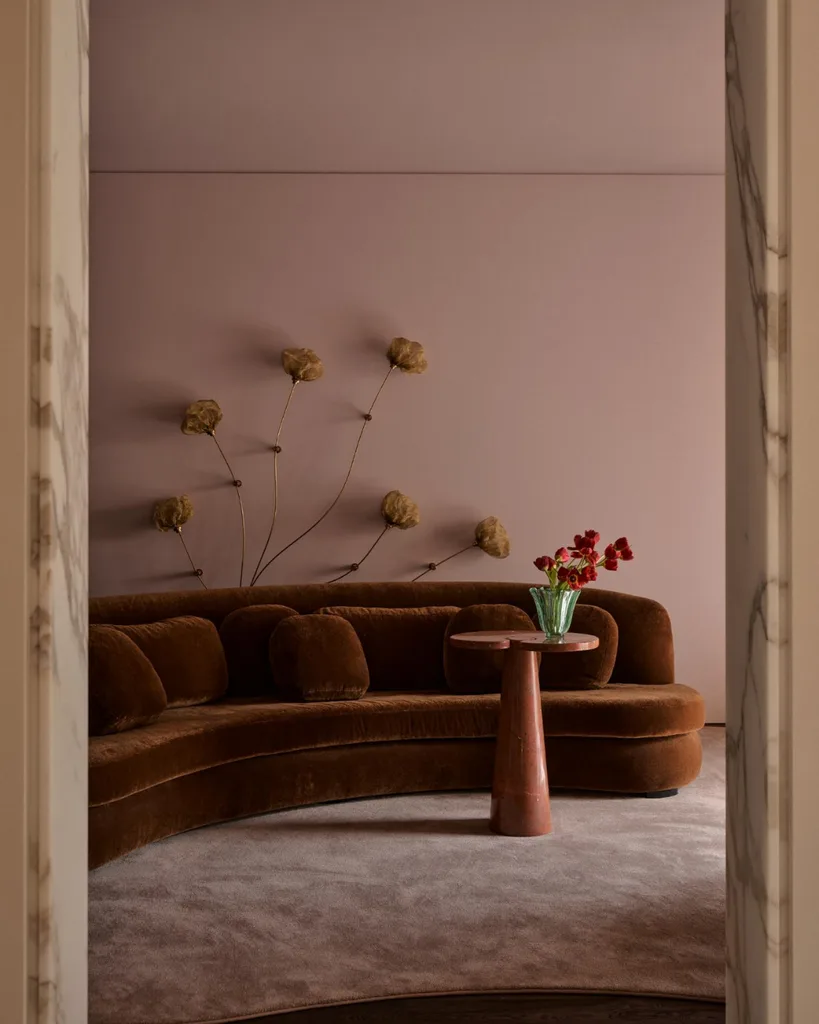 Curved brown sofa with floral wall art, pink walls, and a small table with a vase of red flowers.