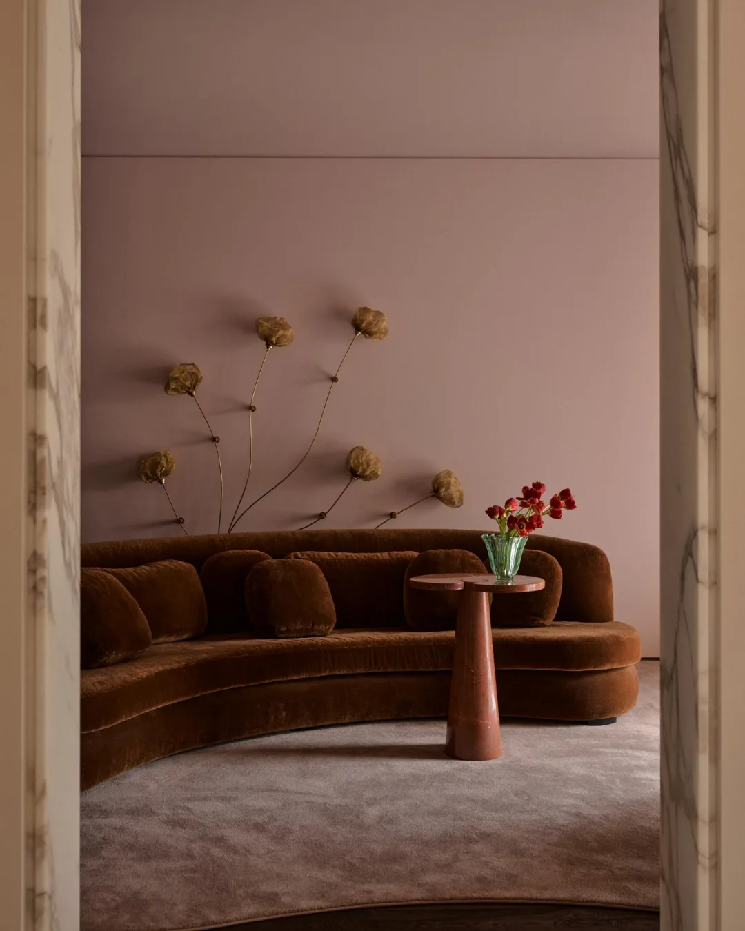 Curved brown sofa with floral wall art, pink walls, and a small table with a vase of red flowers.