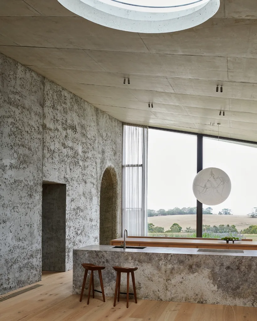 Modern kitchen with stone textures, wooden stools, skylight, large window with scenic view, and a spherical pendant light.