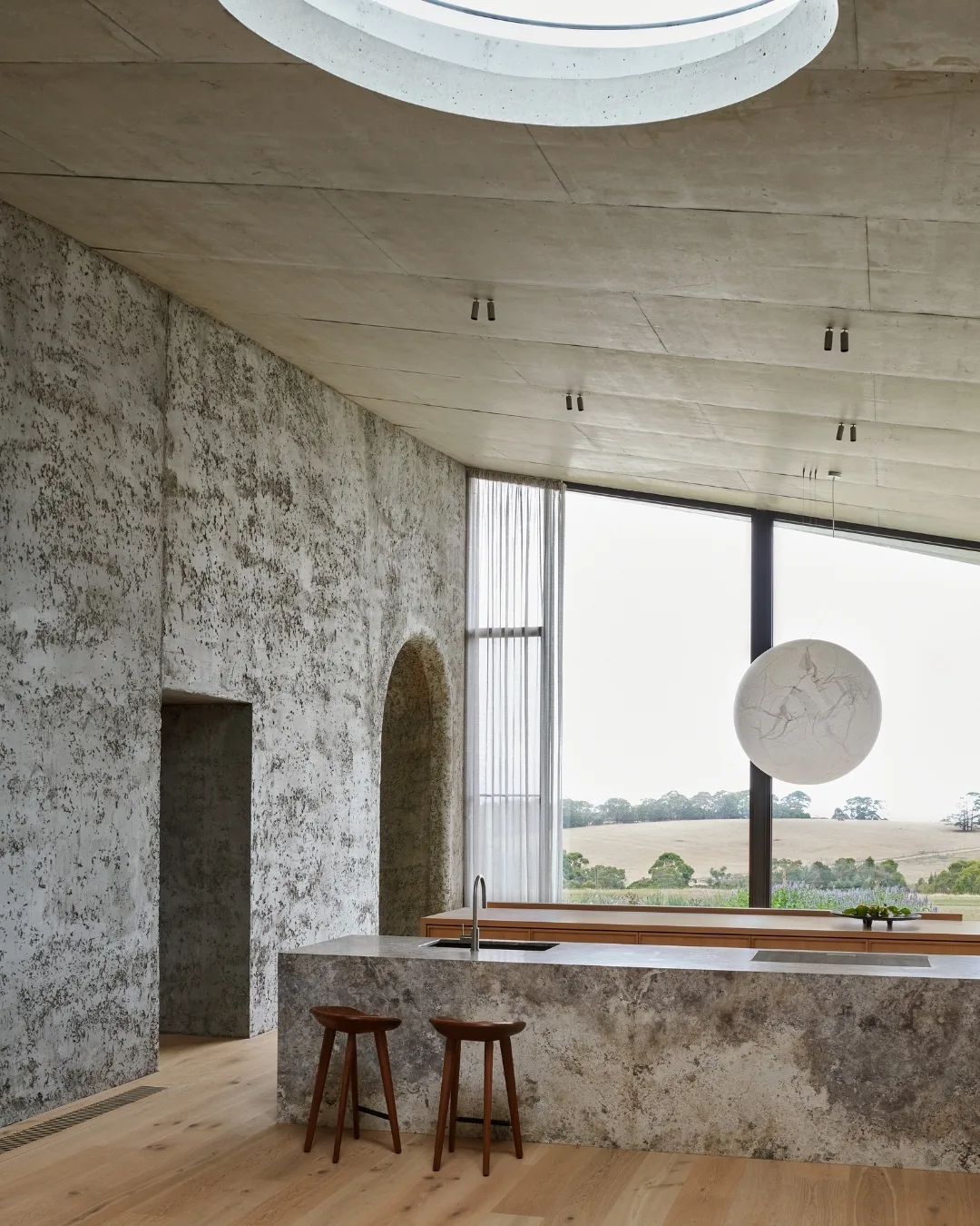 Modern kitchen with stone textures, wooden stools, skylight, large window with scenic view, and a spherical pendant light.	