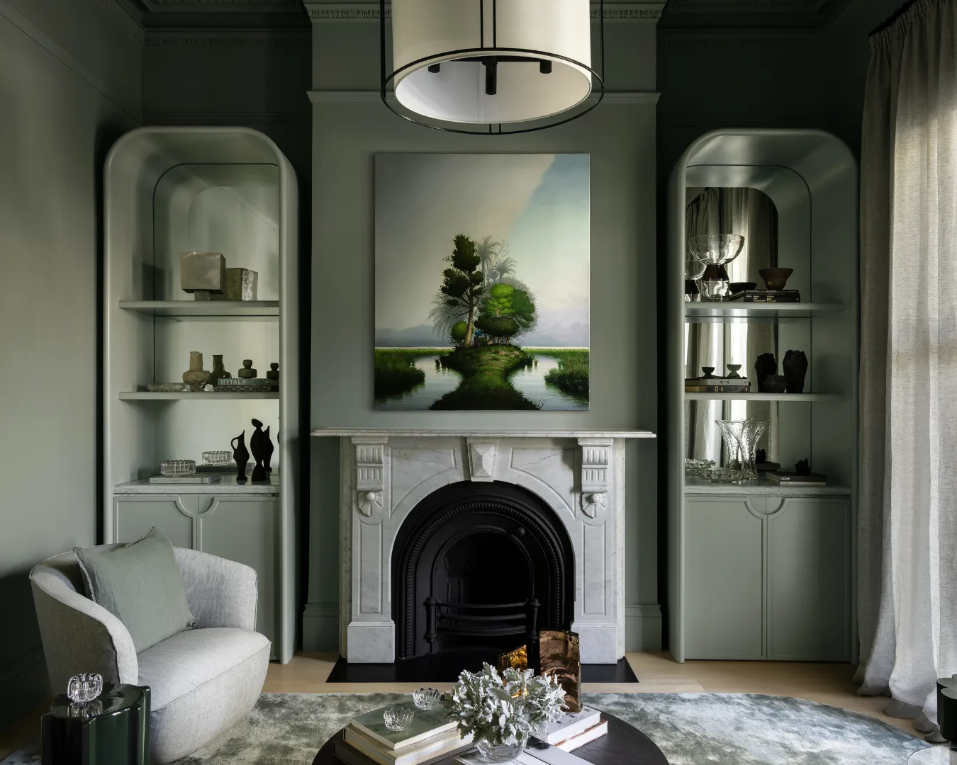 Sophisticated living room with a marble fireplace, artwork, built-in shelves, and soft green-gray walls. Mim Design verdant residence.