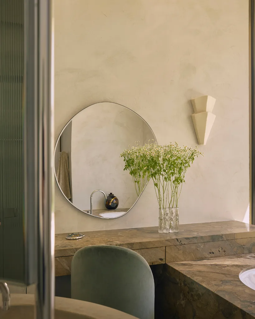 Elegant bathroom with round mirror, marble countertop, and tall flowers in a vase. Soft lighting creates a serene atmosphere.