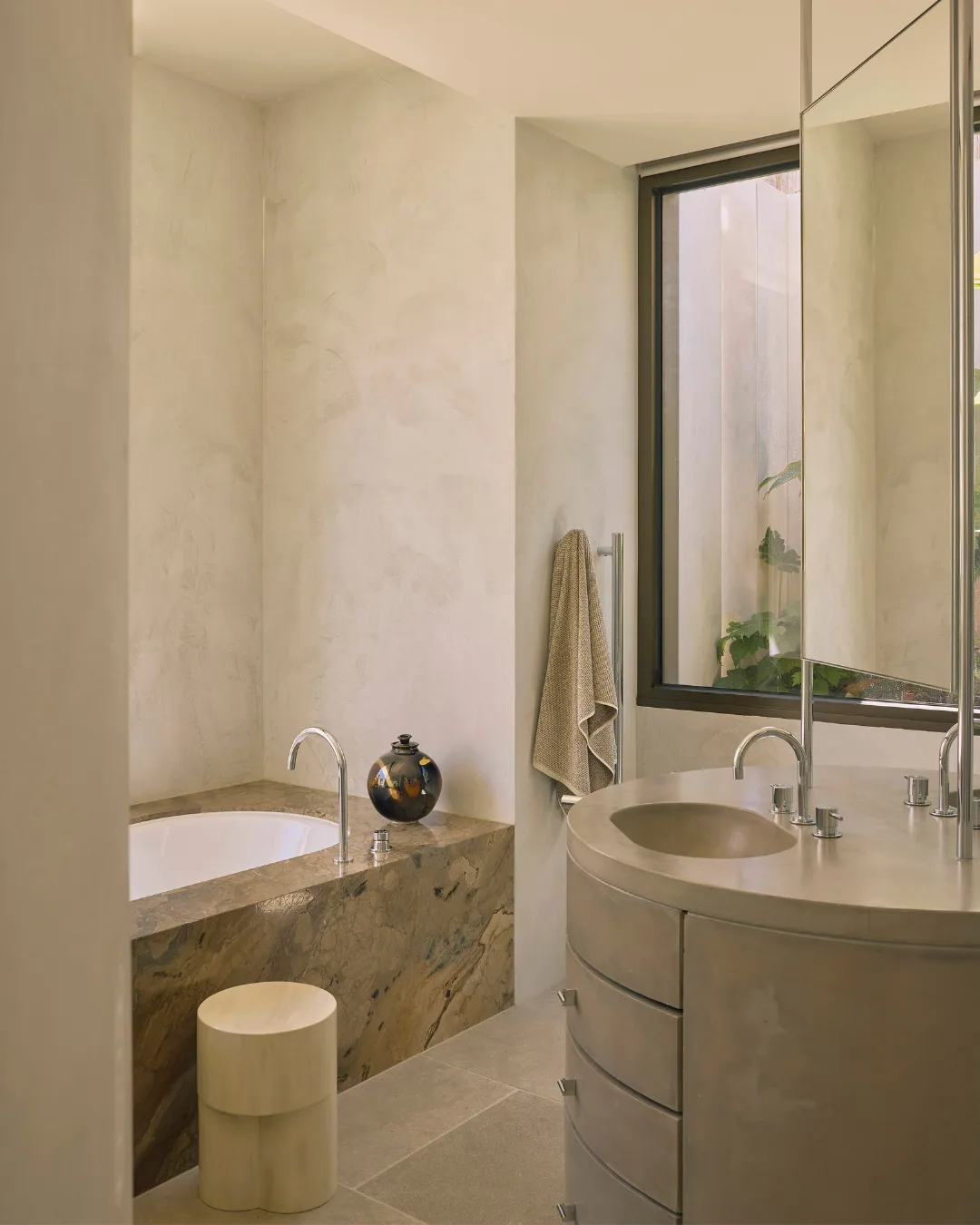 Modern bathroom with a marble bathtub, round sink, stool, and large window. Beige tones and minimalist design.