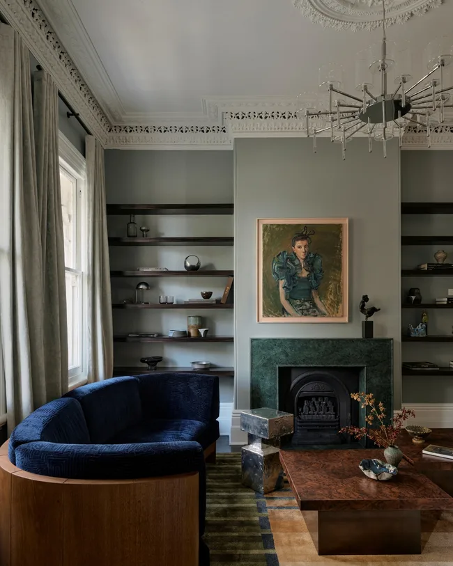 Elegant living room with blue sofa, art above green fireplace, stylish shelves, and modern chandelier.
