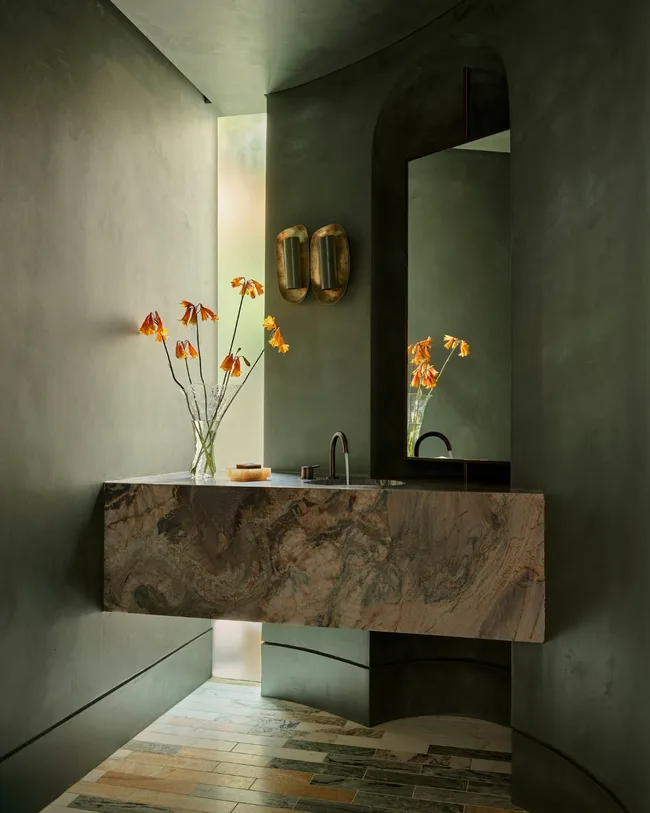 Marble bathroom with green walls, a floating sink, orange flowers in a vase, and a large mirror.