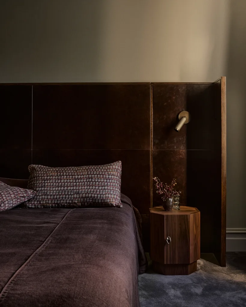 Dark, cozy bedroom with a brown bed, patterned pillow, wooden nightstand, and decorative flowers.