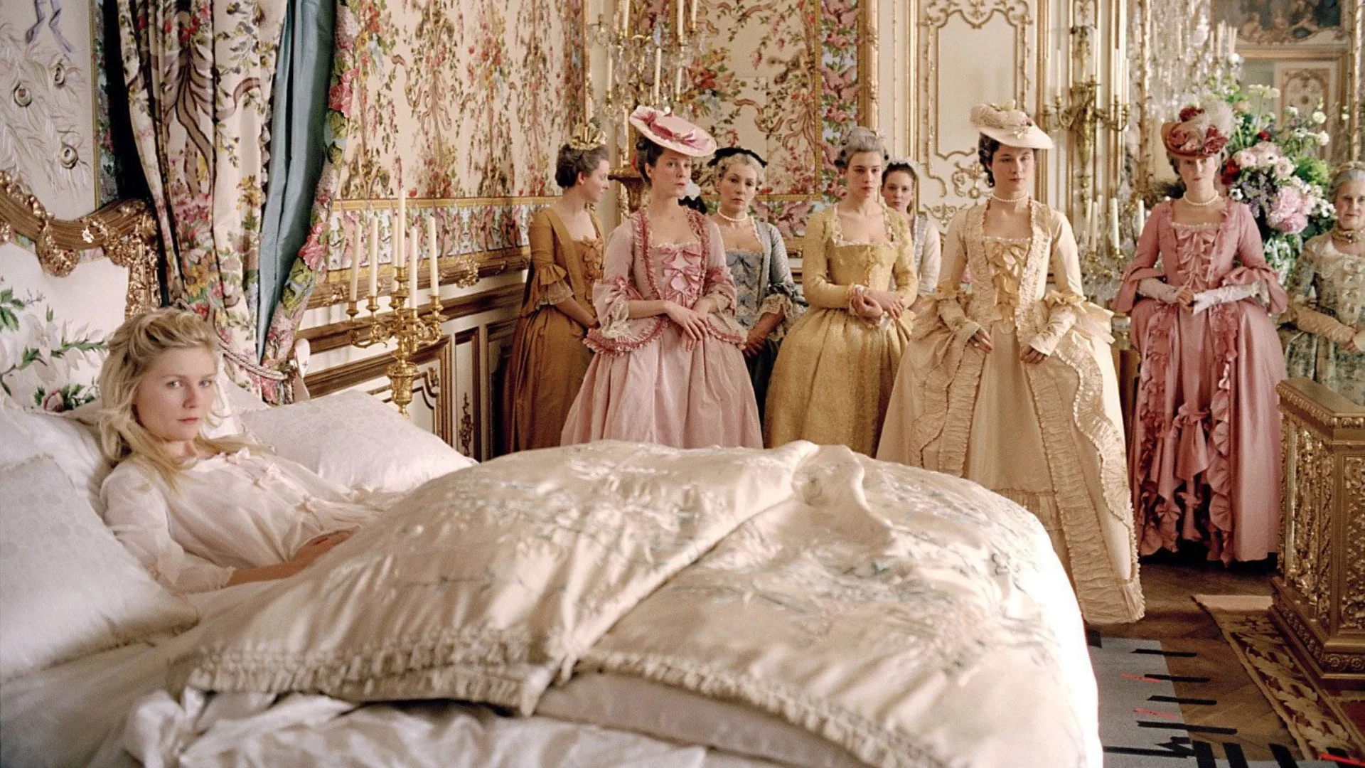 A still image from the Sofia Coppola film 'Marie Antoinette' showing Kirsten Dunst's character in a room with decorative floral wallpaper, gilded candles and mouldings and a group of women dressed in regal attire. 