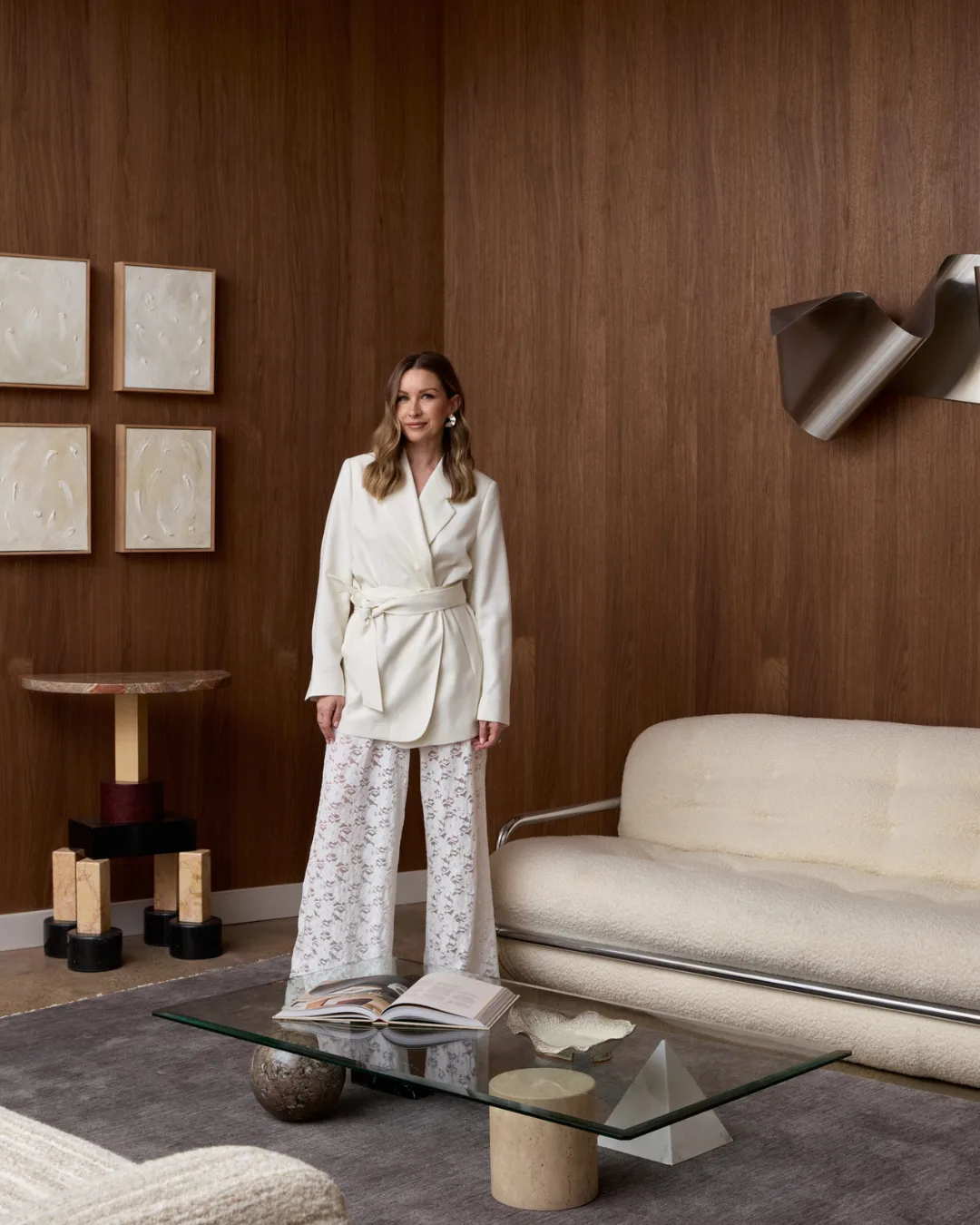 Woman in white pantsuit standing in front of boucle couch, glass coffee table and walnut timber walls.