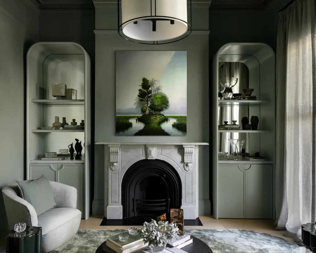 Elegant green living room with fireplace, modern art above, and shelves with decor. Cozy chair and soft lighting enhance the space.
