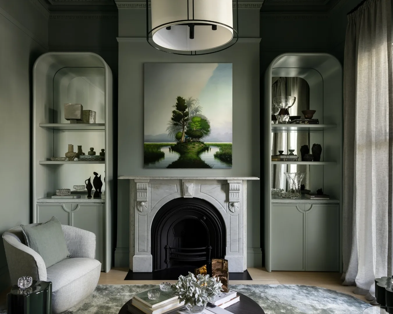 Elegant green living room with fireplace, modern art above, and shelves with decor. Cozy chair and soft lighting enhance the space.