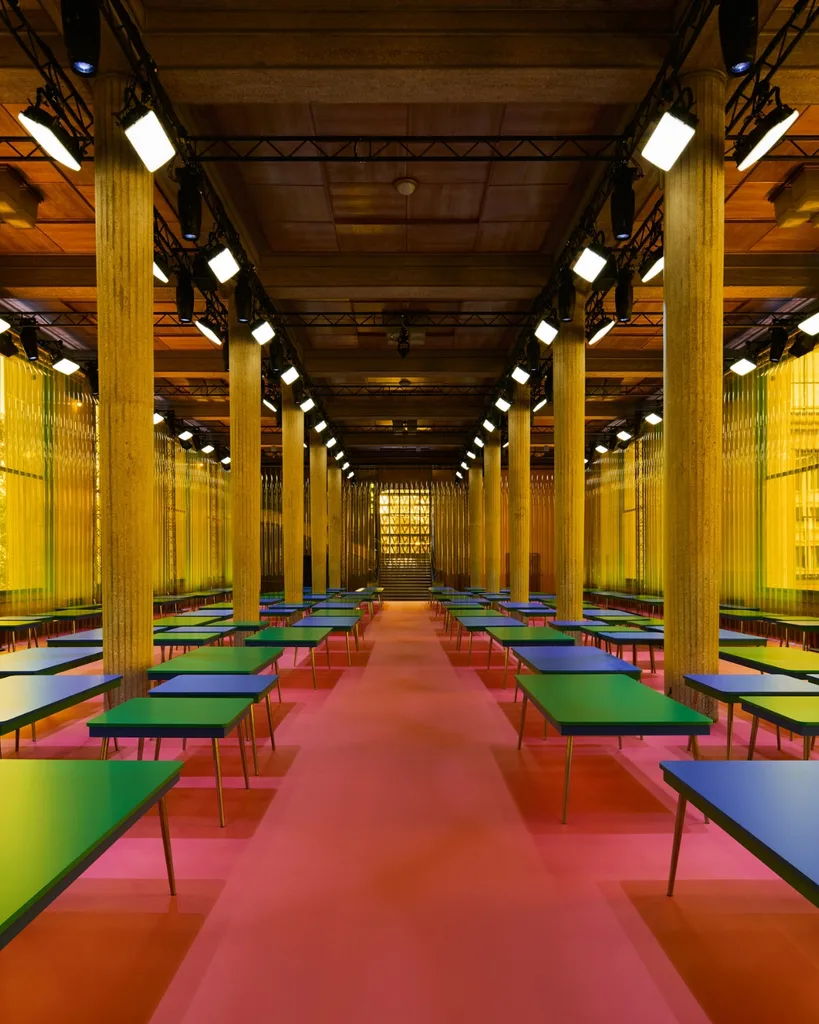 Miu Miu fashion month Spring Summer 2026 show set design features a room with rows of Formica tables in primary colours on a red floor surrounded by yellow walls of the Palais d’Iéna.