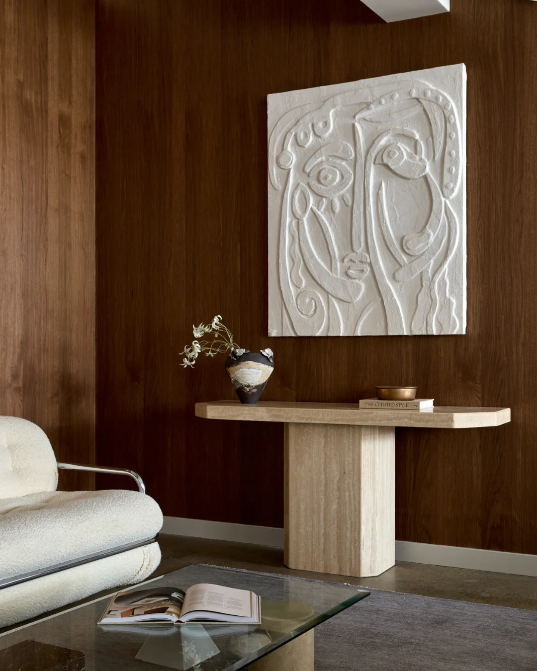 Ceramic sideboard with charcoal, ceramic vase filled with flowers, coffee table book, white contemporary artwork of face, glass coffee table and cream, boucle couch.