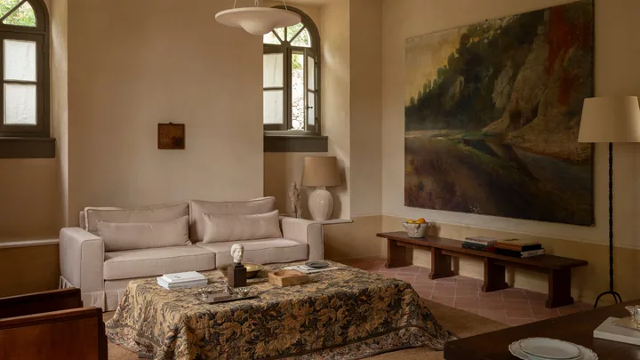 A living room with a grey linen couch, artwork of natural landscape, pattern fabric coffee table, terracotta brickwork flooring and frosted arched windows with grey frames.
