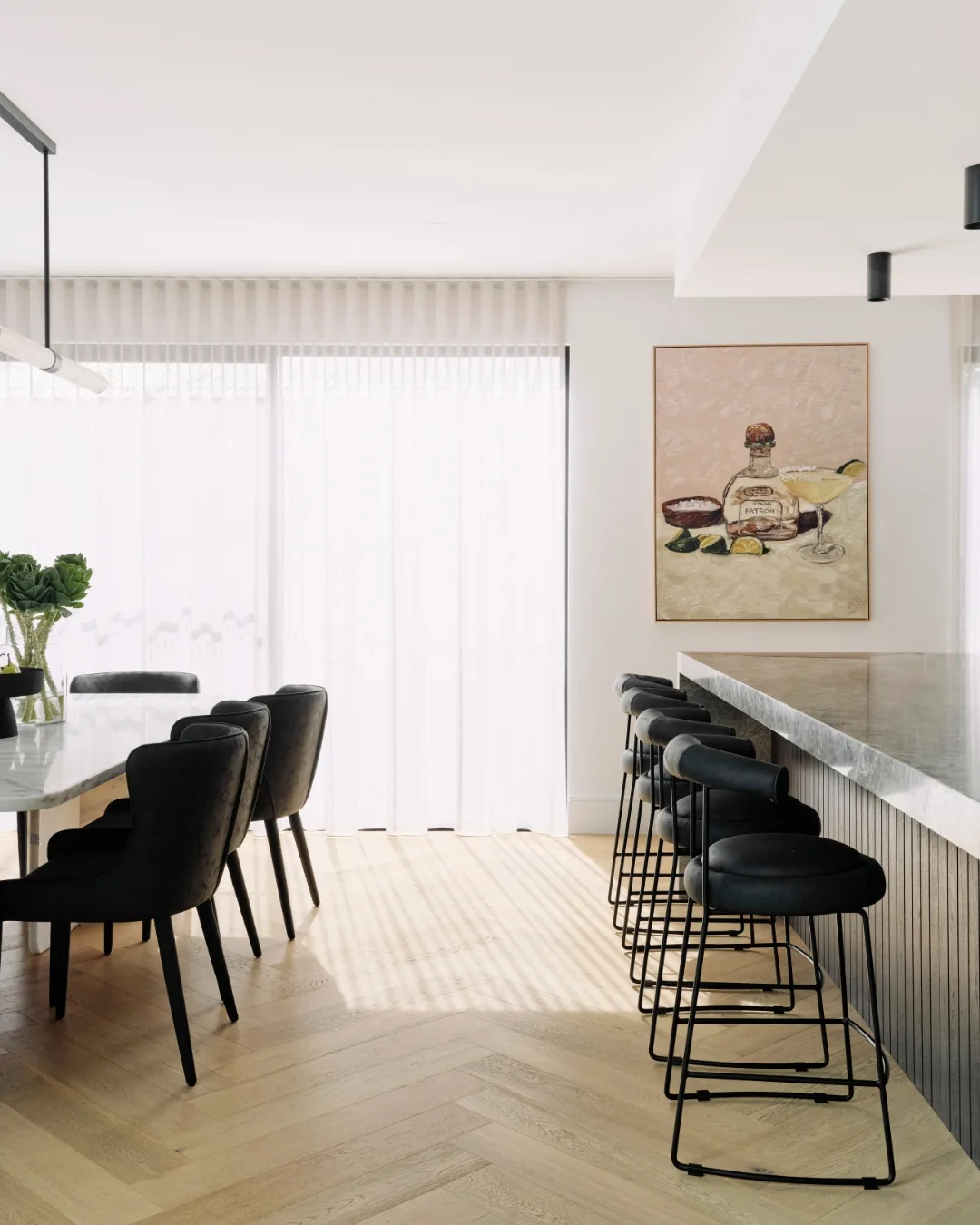 A galley dining room with barstools on the right and dining chairs on the left, with herringbone engineered timber flooring, looking out to a light window covered in sheer curtains.