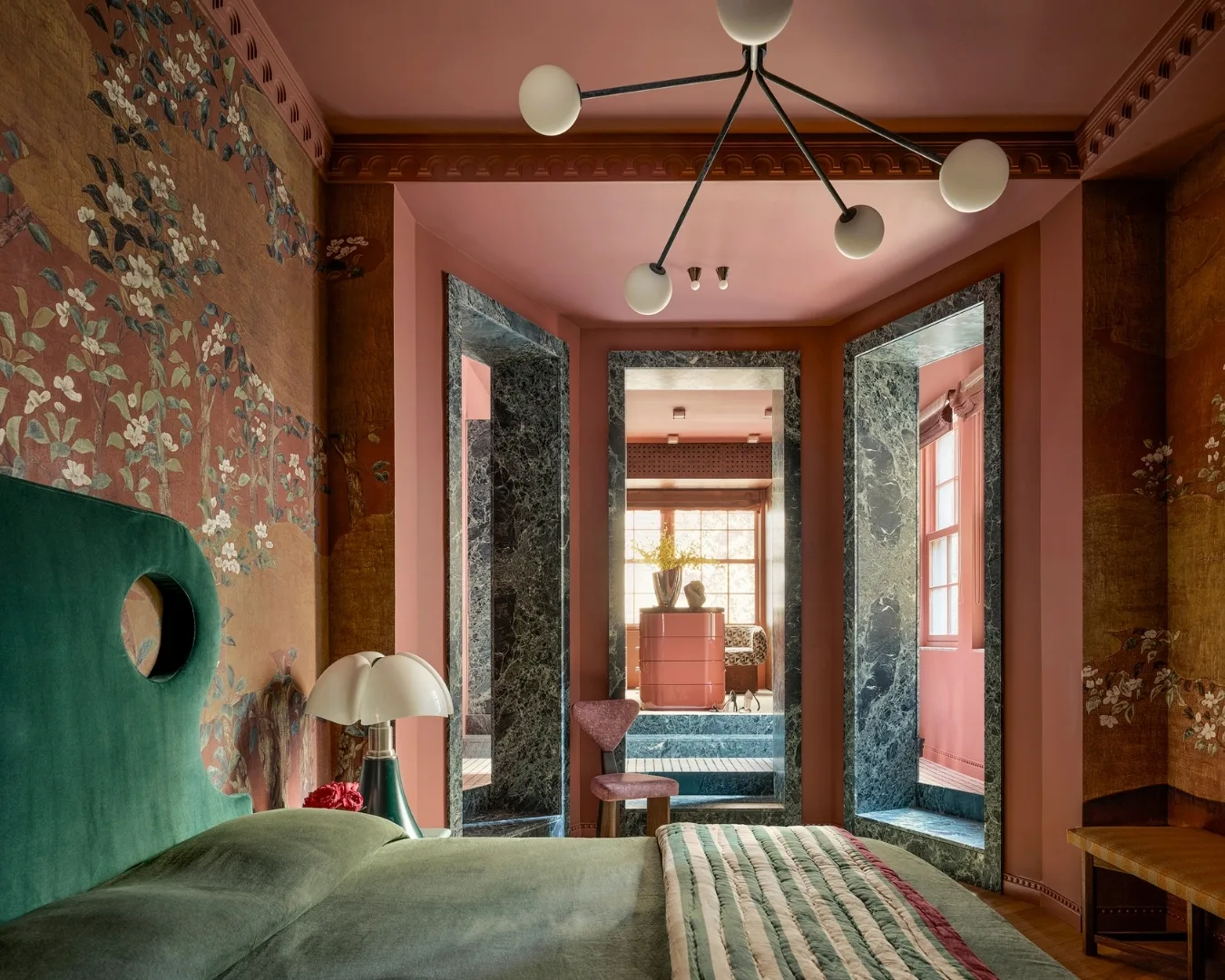 Cozy bedroom with floral wallpaper, green headboard, unique chandelier, and a view into an adjoining room with large windows. 