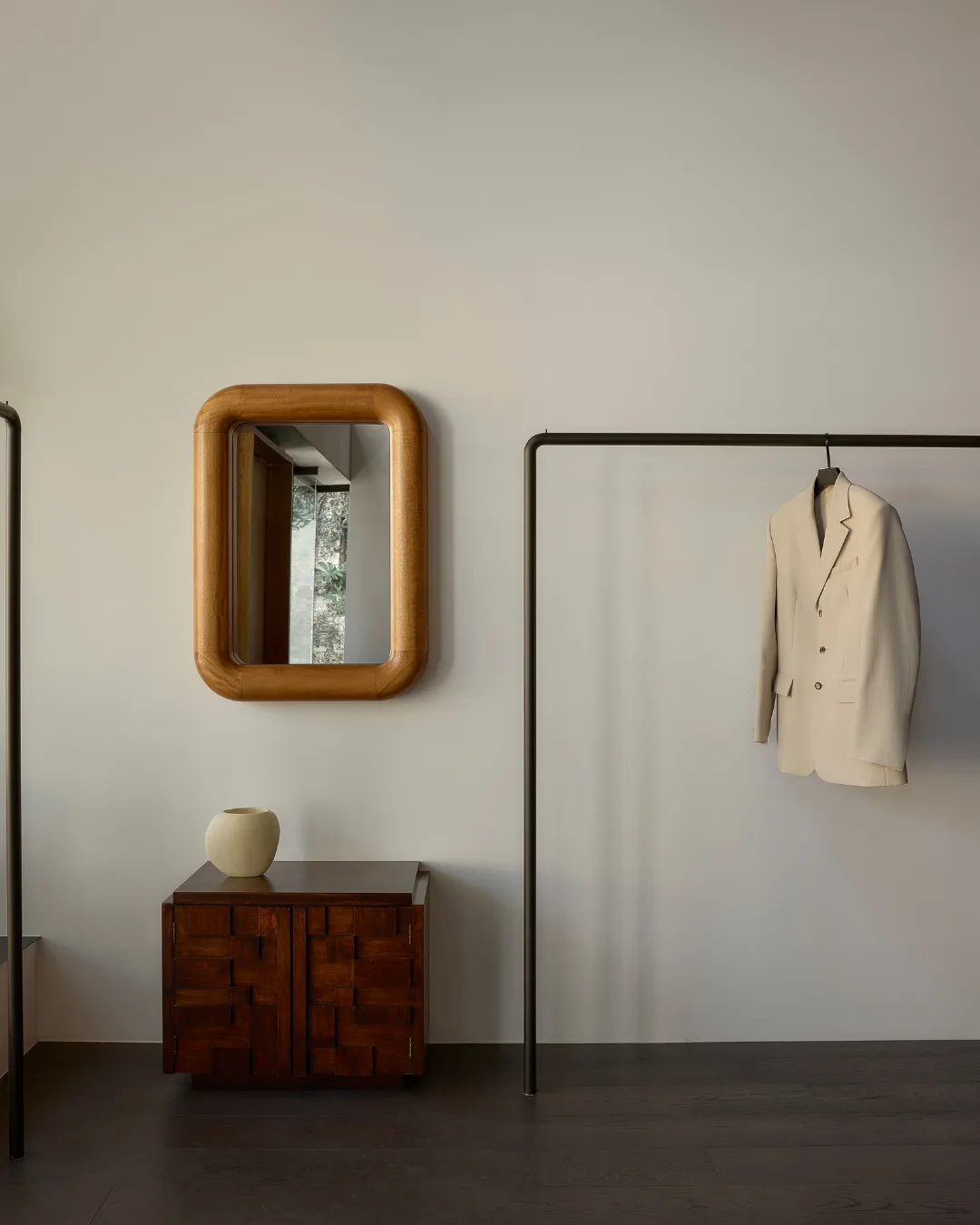 Preview Mirror with wooden frame above a cabinet and vase; beige suit jacket on a clothes rack in minimalist interior.