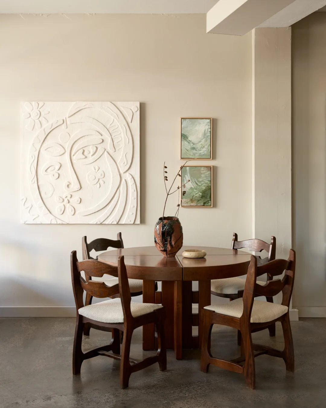 Wooden dining table and chairs with cream boucle cushion covers, ceramic vase and surrealist paintings hanging on the wall.