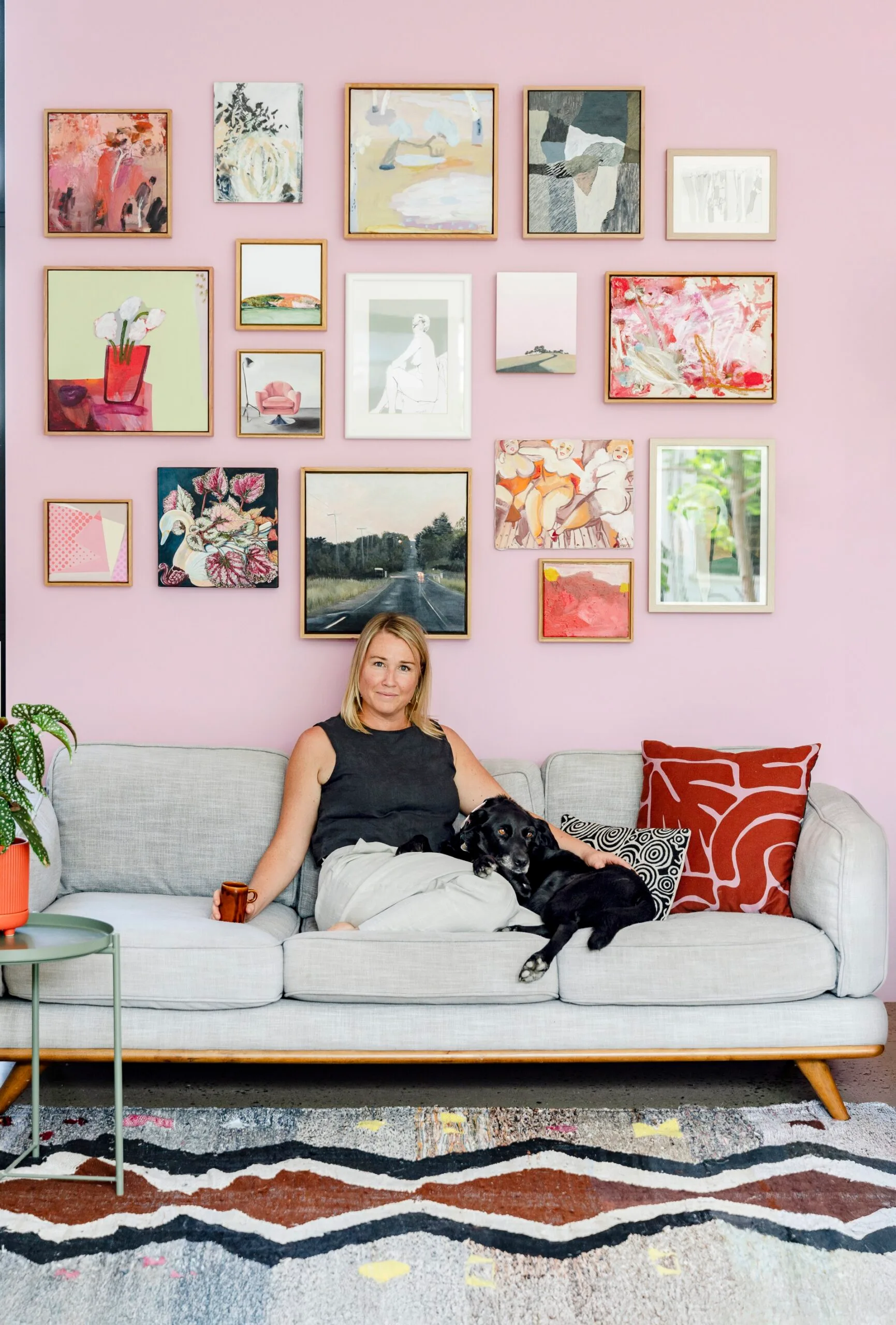 Artist Madi Young sits on a grey sofa beneath a gallery wall. Her dog lays beside her.