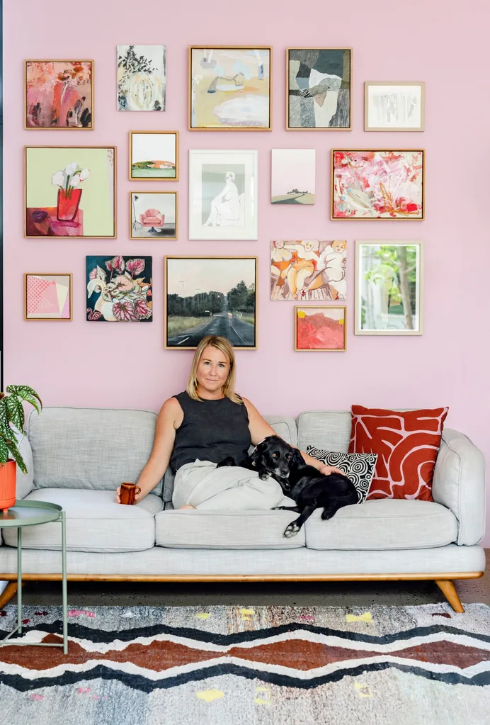 Artist Madi Young sits on a grey sofa beneath a gallery wall. Her dog lays beside her.