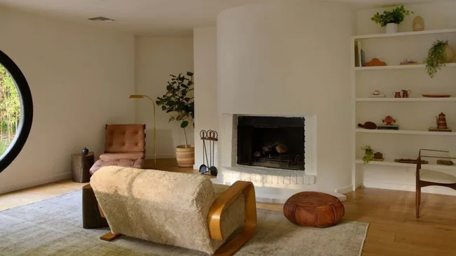 a living room with white fireplace, beige couch, leather footstool and bookshelf