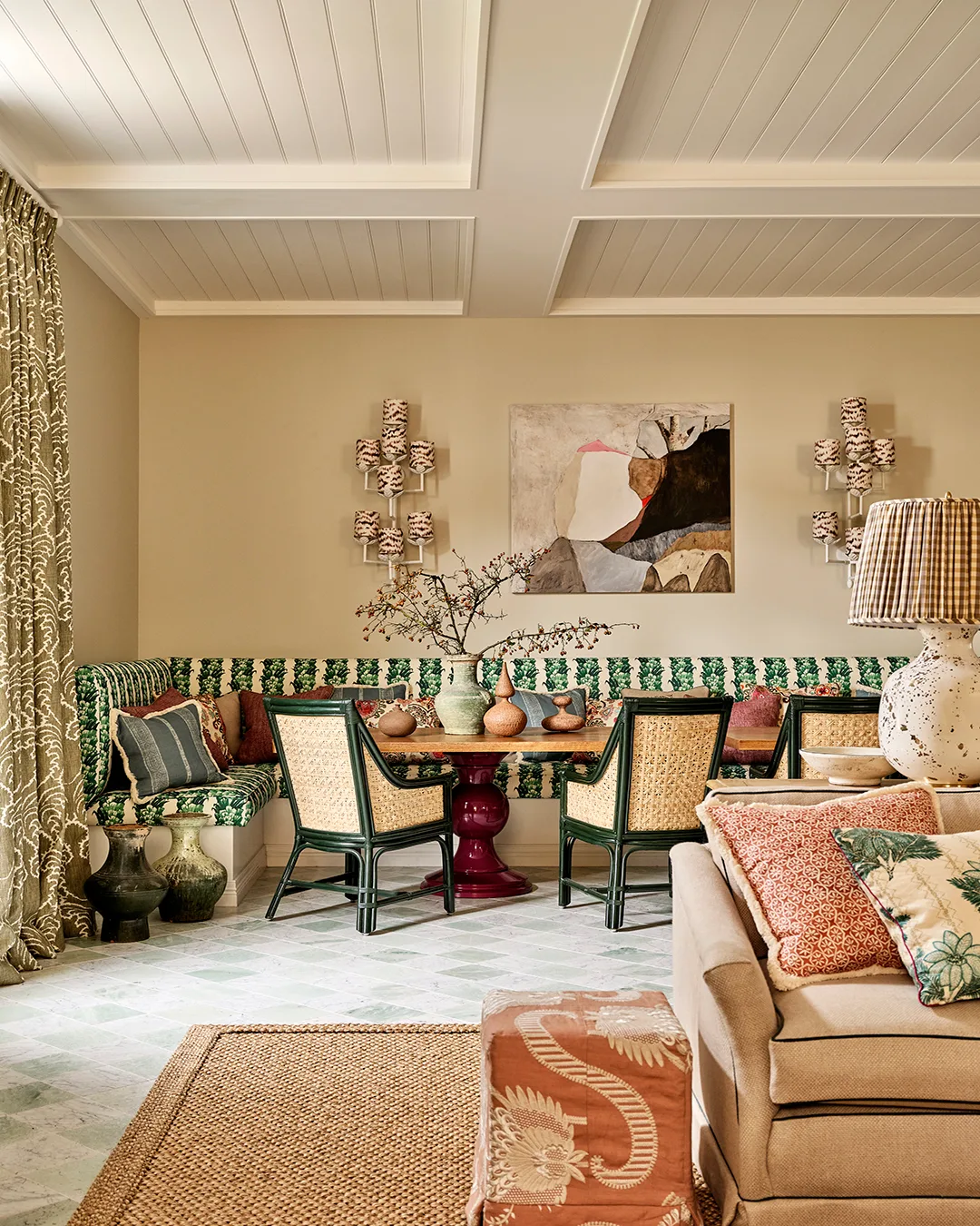 A dining room with rattan chairs, green pattern sofa, abstract art and beige couch with coloured cushions