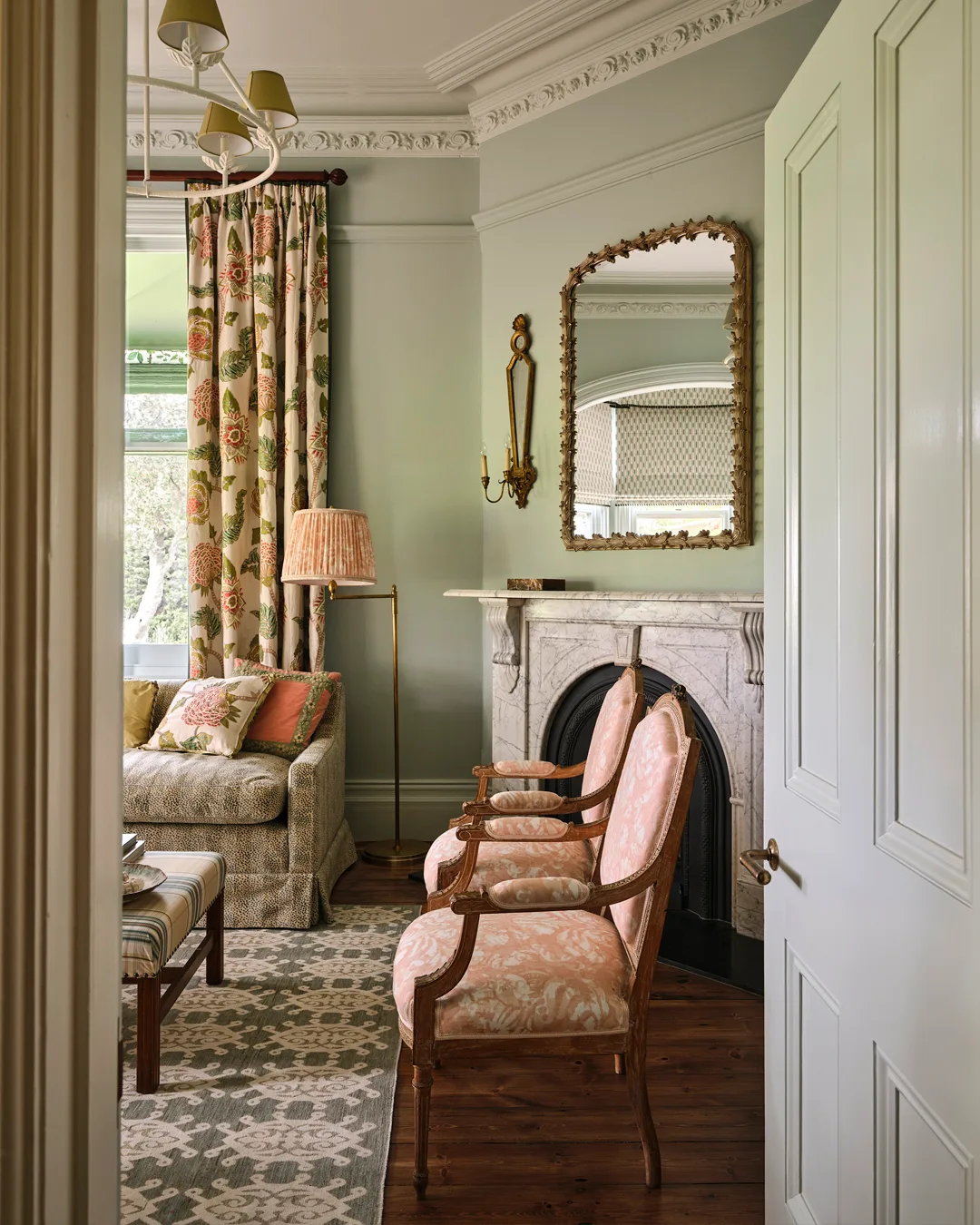 A living room with green walls, pink pattern chairs, mirror and fireplace painted white