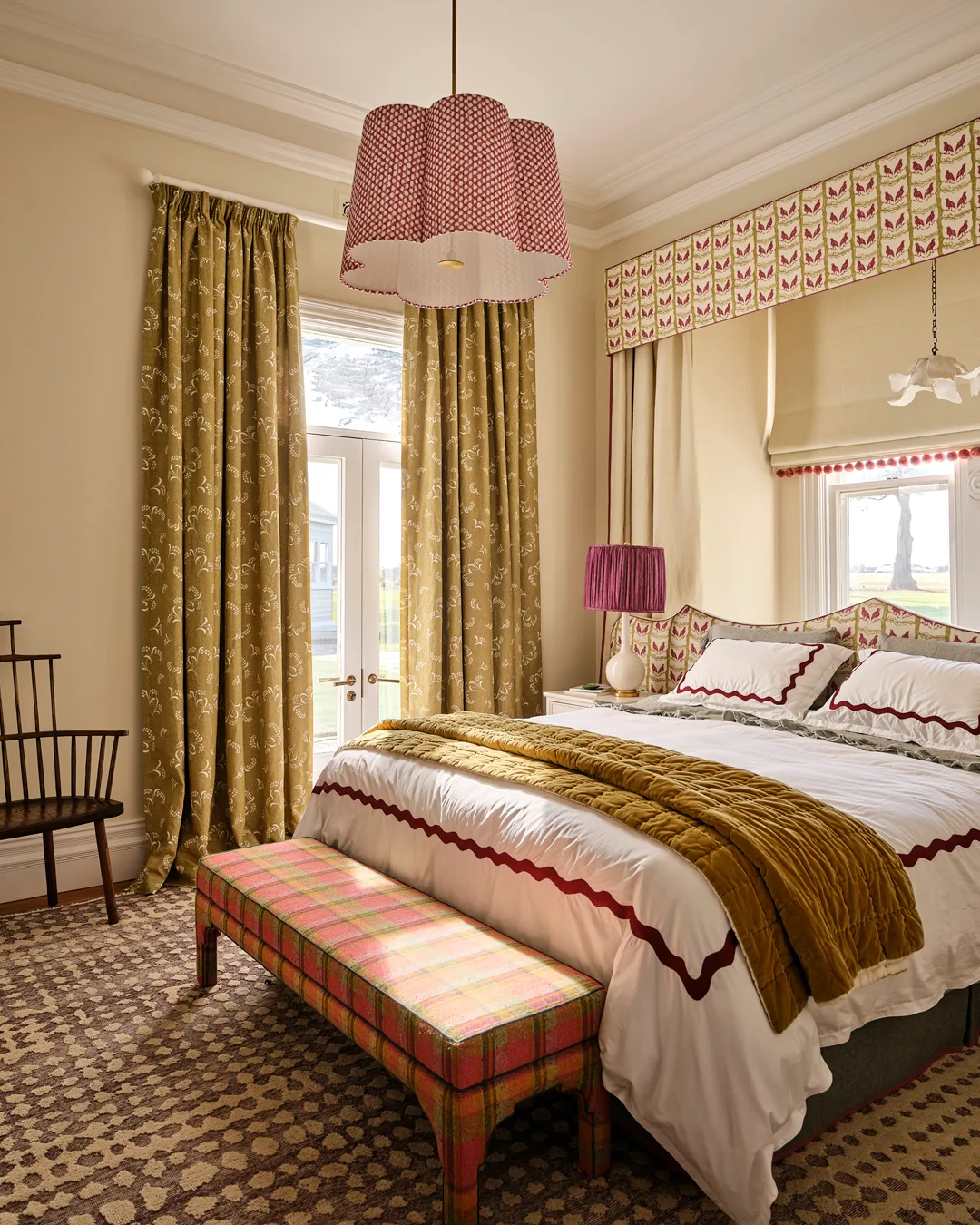 A bedroom with mustard pattern curtains, bed with white linen and pink embroidery, pink pendant lampshade, timber chair and mustard throw
