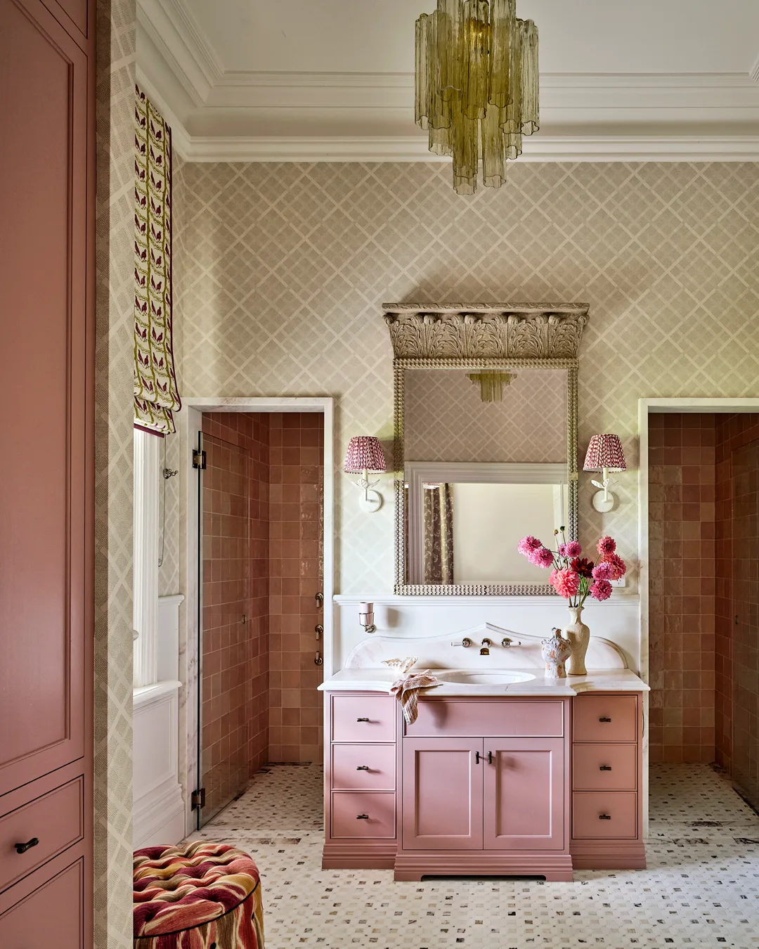 A bathroom with pink cabinetry scalloped mirror, gold chandelier, pink tiled shower and pink flowers.