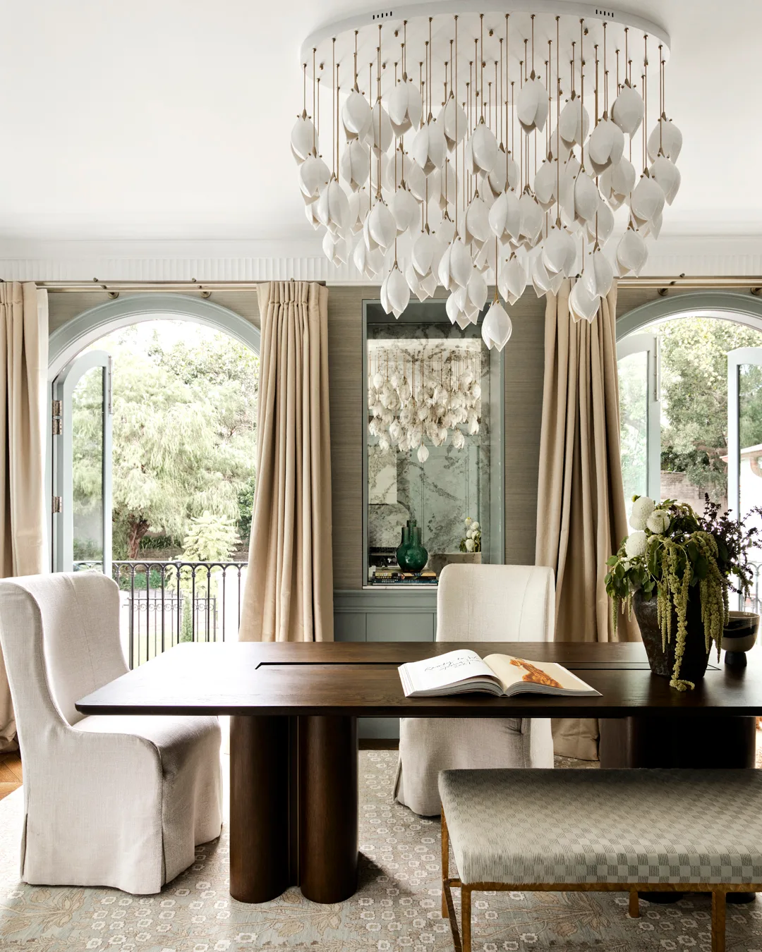 A timber dining table with plush cream chairs, white modern chandelier, beige curtains, grey joinery, and grey, textured wallpaper